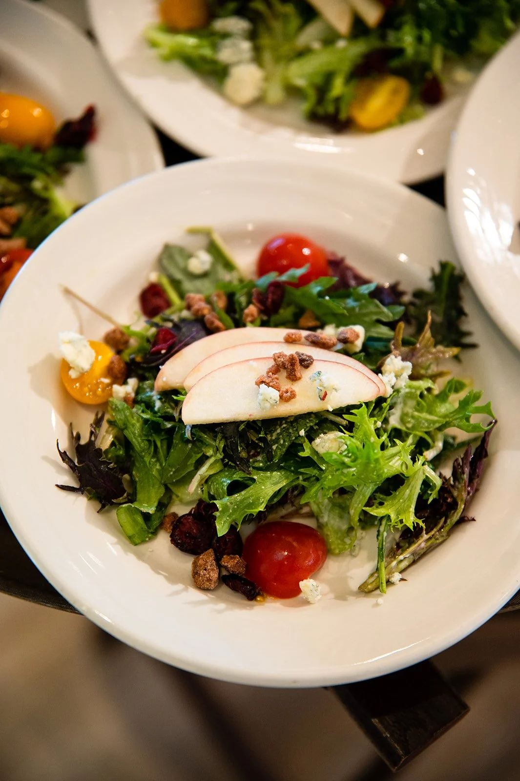 A fresh mixed salad with cherry tomatoes, sliced apples, crumbled blue cheese, and candied pecans on a white plate.