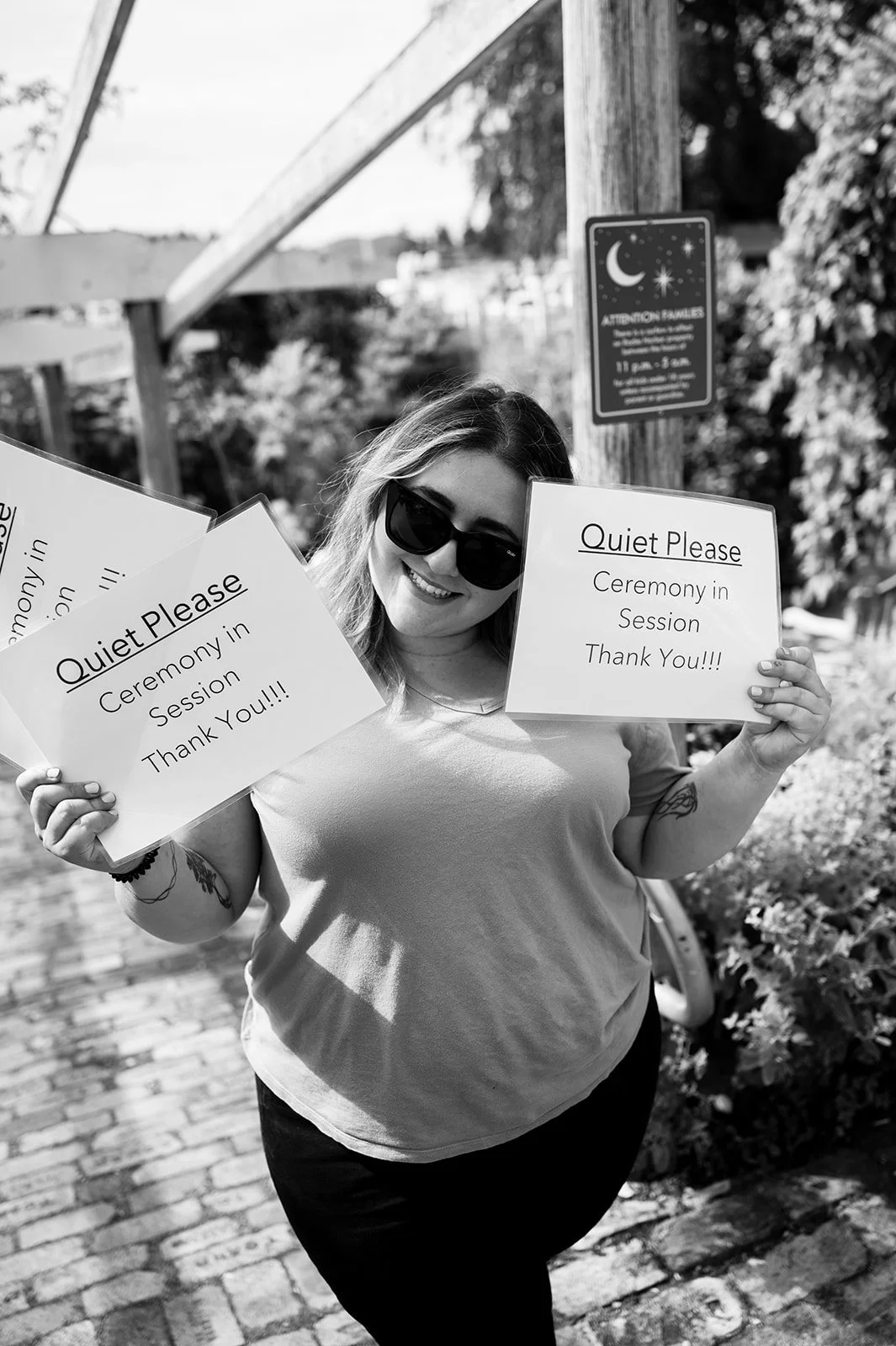 Smiling woman with sunglasses holding signs that read 'Quiet Please Ceremony in Session Thank You!!!' in an outdoor setting.