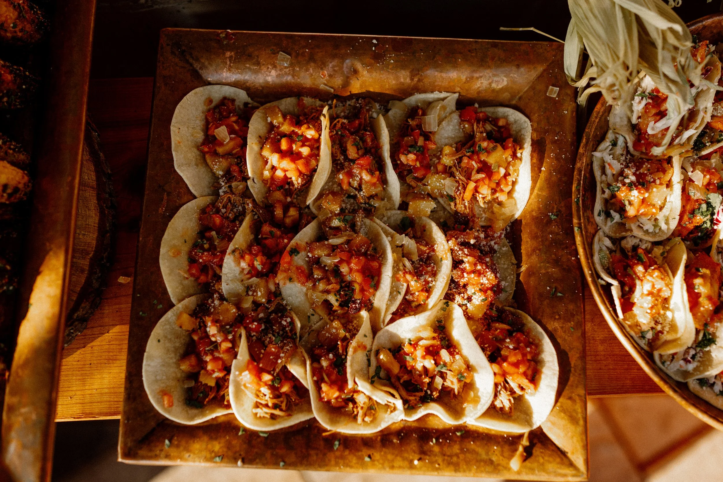 Close-up of a tray of tacos filled with diced vegetables and sauce, with another plate of tacos partially visible on the right side.