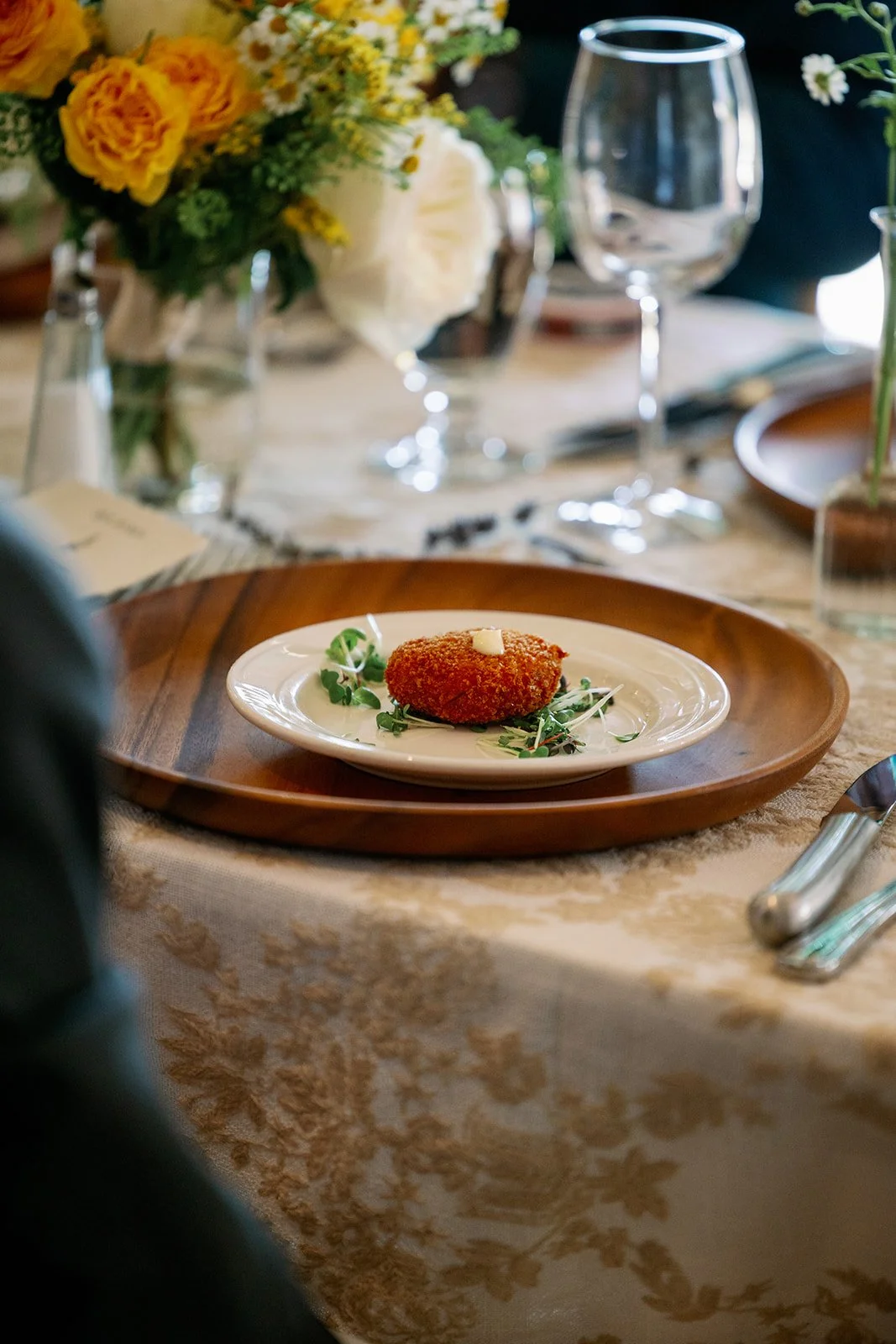 A plated piece of fried food garnished with microgreens on a white plate, set on a wooden charger on a decorated dining table.