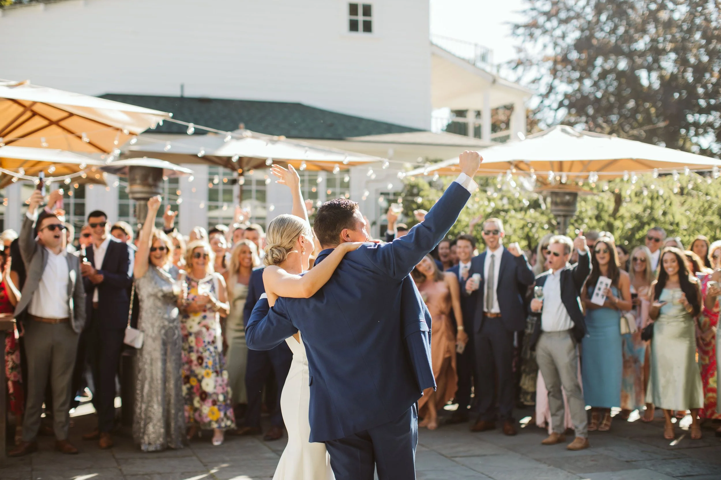 A newlywed couple dancing at their outdoor wedding reception with their wedding guests cheering and celebrating in the background.