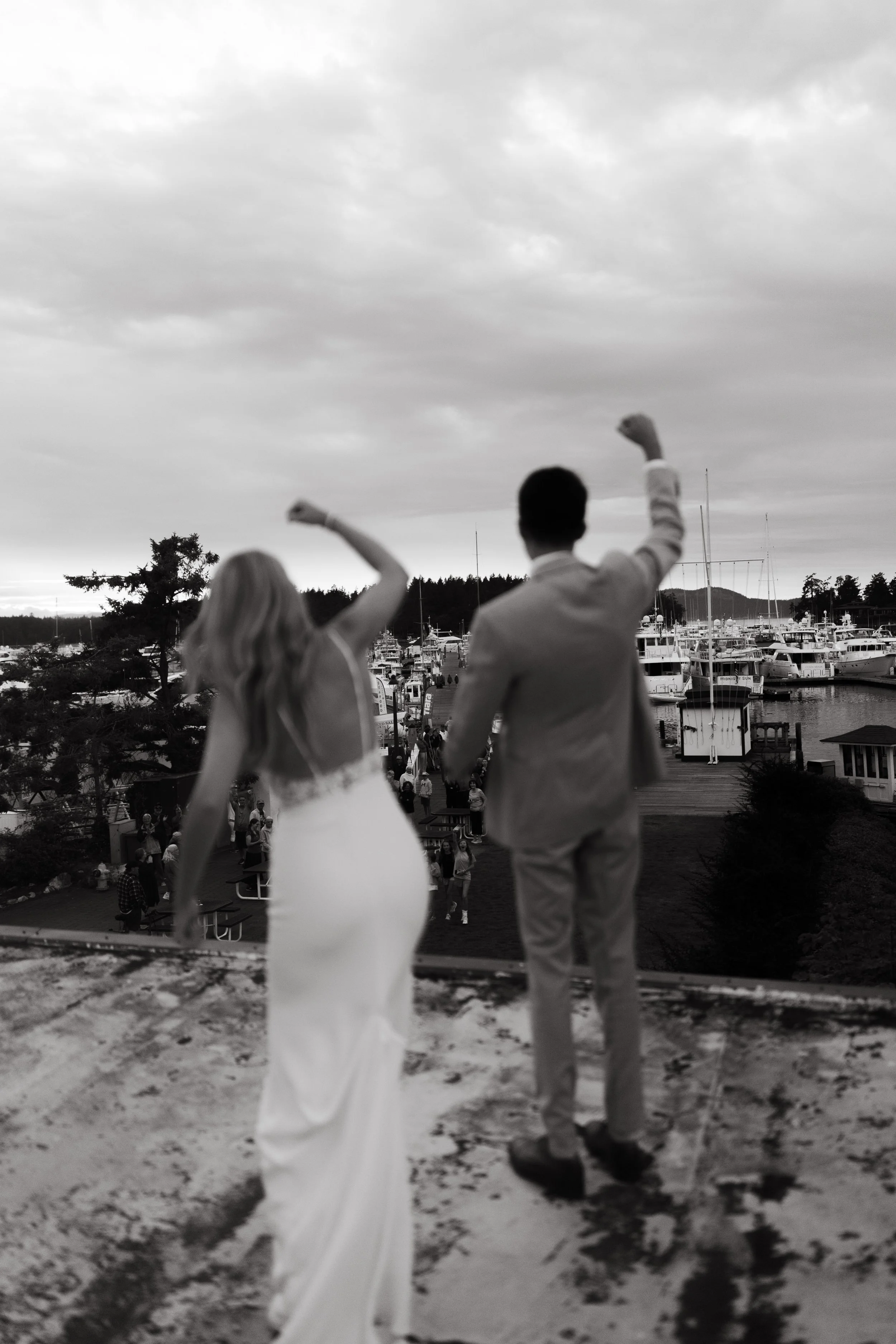 Black and white photo of a bride and groom celebrating on a rooftop overlooking a marina with boats, people, and trees in the background.