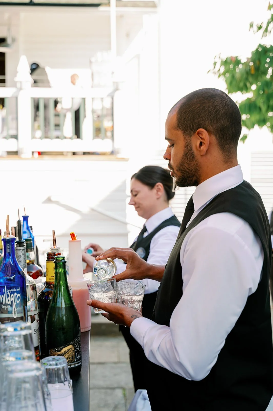 Bartenders preparing drinks outdoors with bottles and glasses on a bar counter.