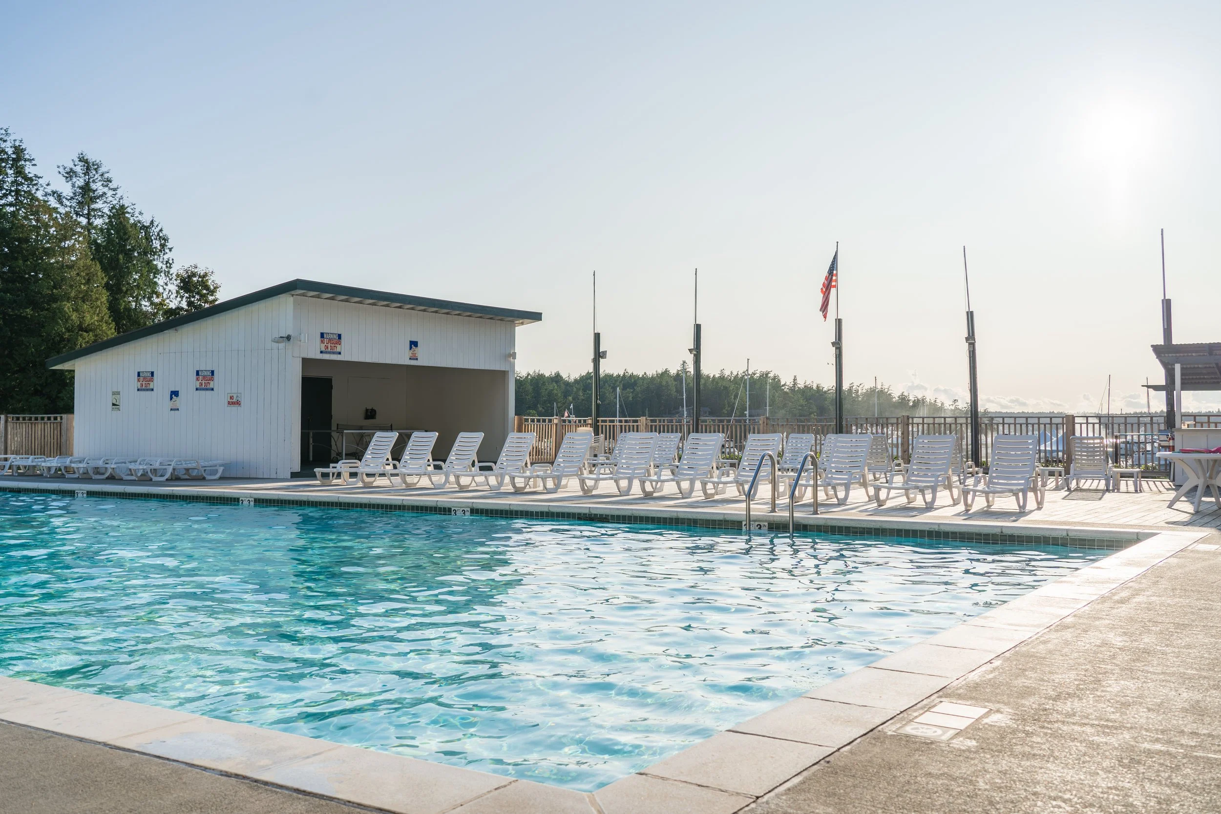 An outdoor swimming pool with several white lounge chairs arranged along the deck, a small white building with signs and flags, and a chain-link fence surrounding the area. The sun is shining brightly in the sky.