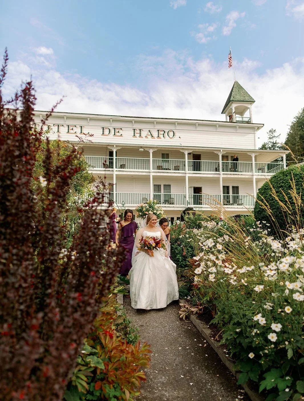A bride in a white wedding dress holding a bouquet, walking with her bridesmaids in purple dresses along a garden path outside a white hotel building with multiple balconies.