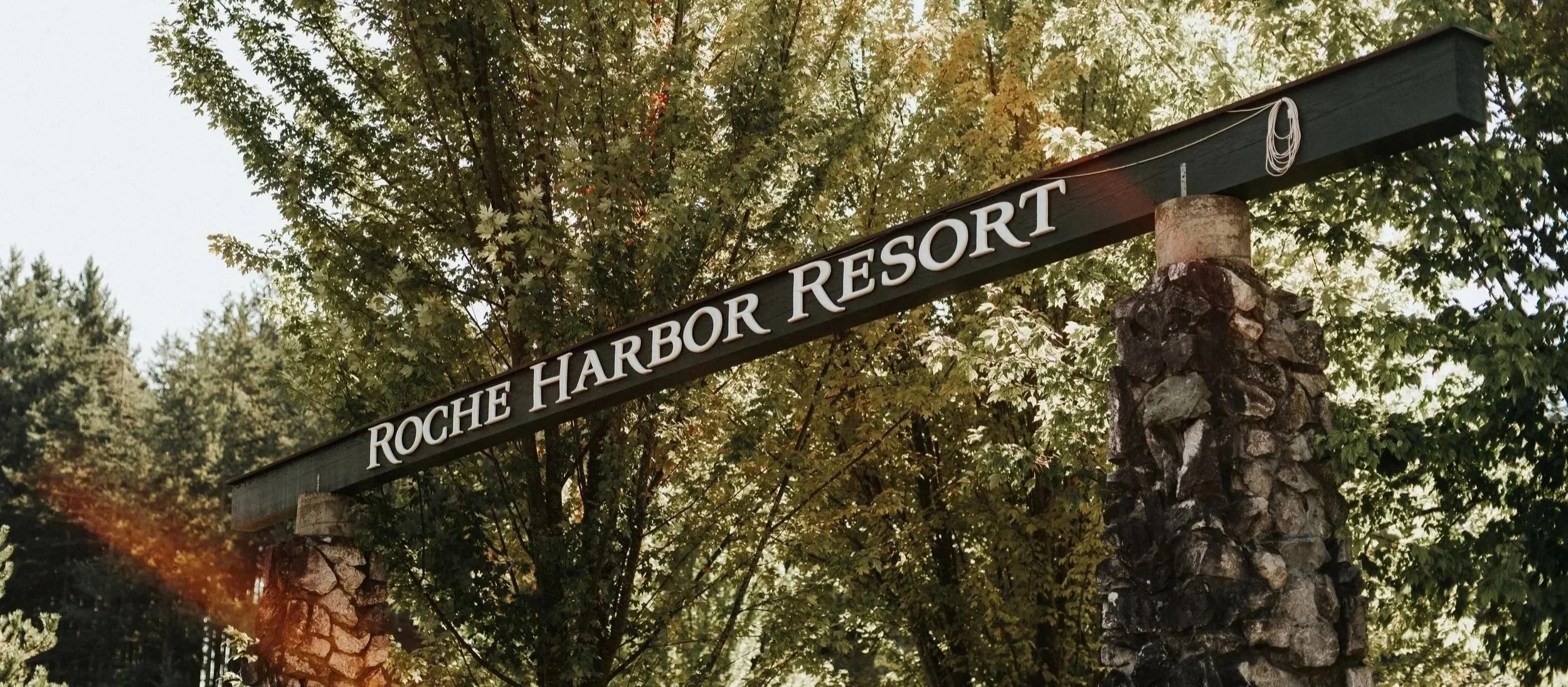 A black horizontal sign with white lettering that reads 'Roche Harbor Resort' is mounted between two stone pillars, with trees in the background.