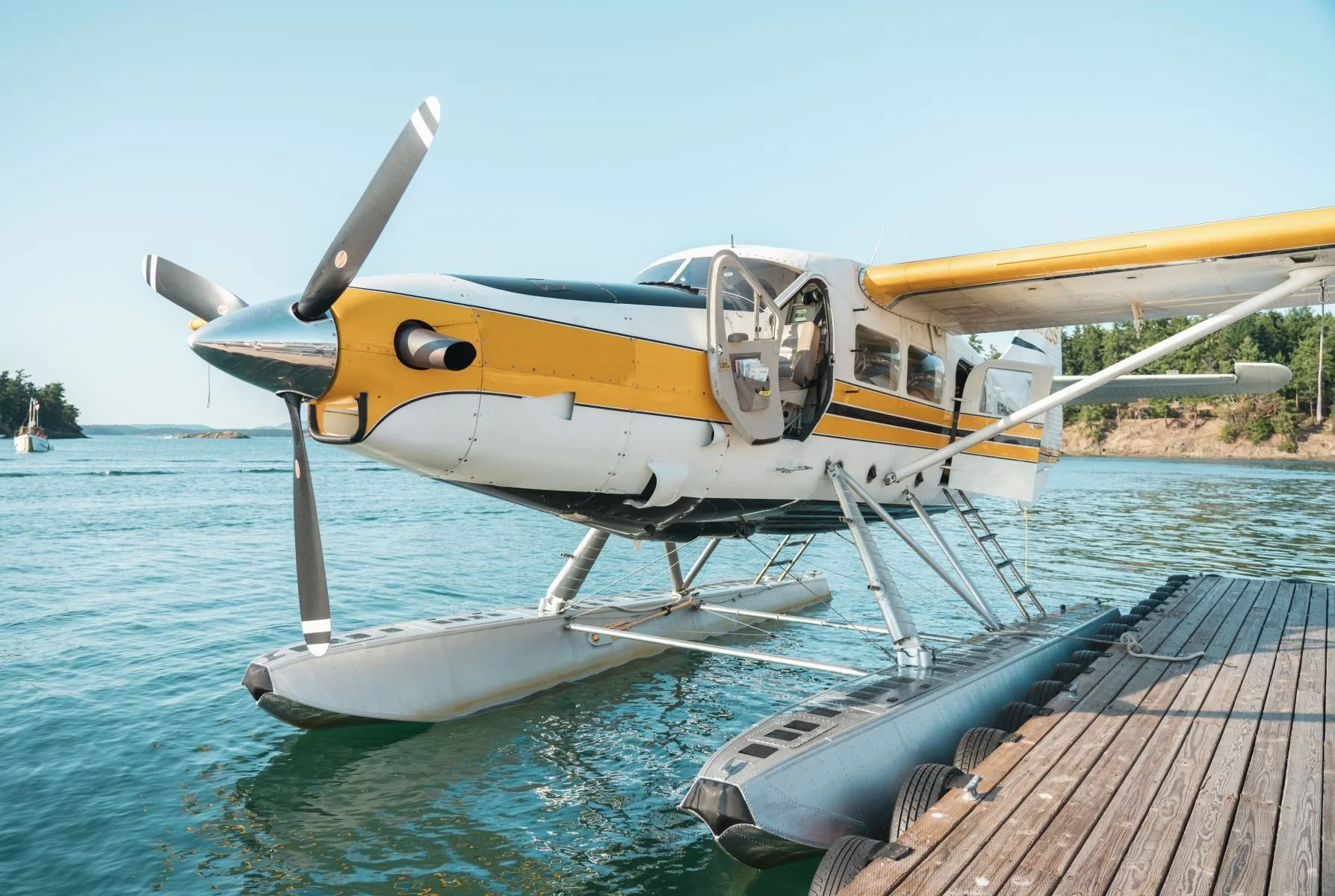 Small seaplane docked at a wooden pier on a clear day with calm water and a distant shoreline with trees.