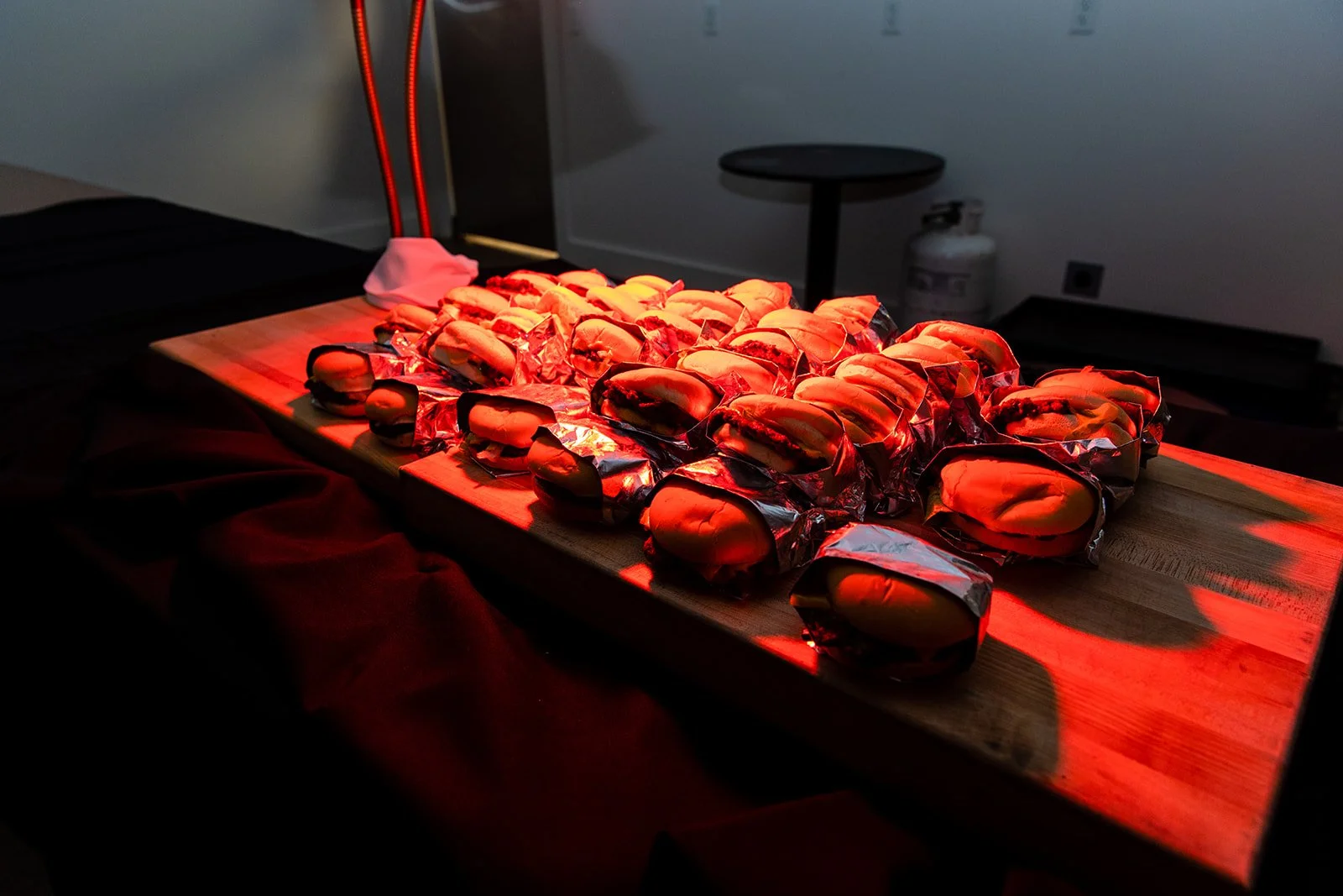 Multiple hot dogs wrapped in aluminum foil sitting on a wooden cutting board, illuminated by red heat lamps in a dark room.