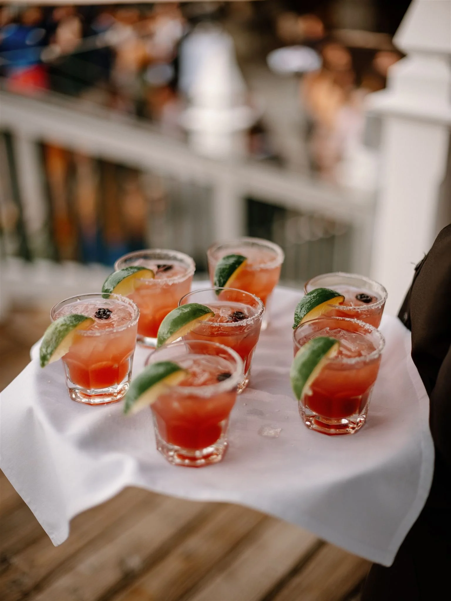 A tray of seven cocktails with lime wedges and black cherries garnish, set on a white napkin.