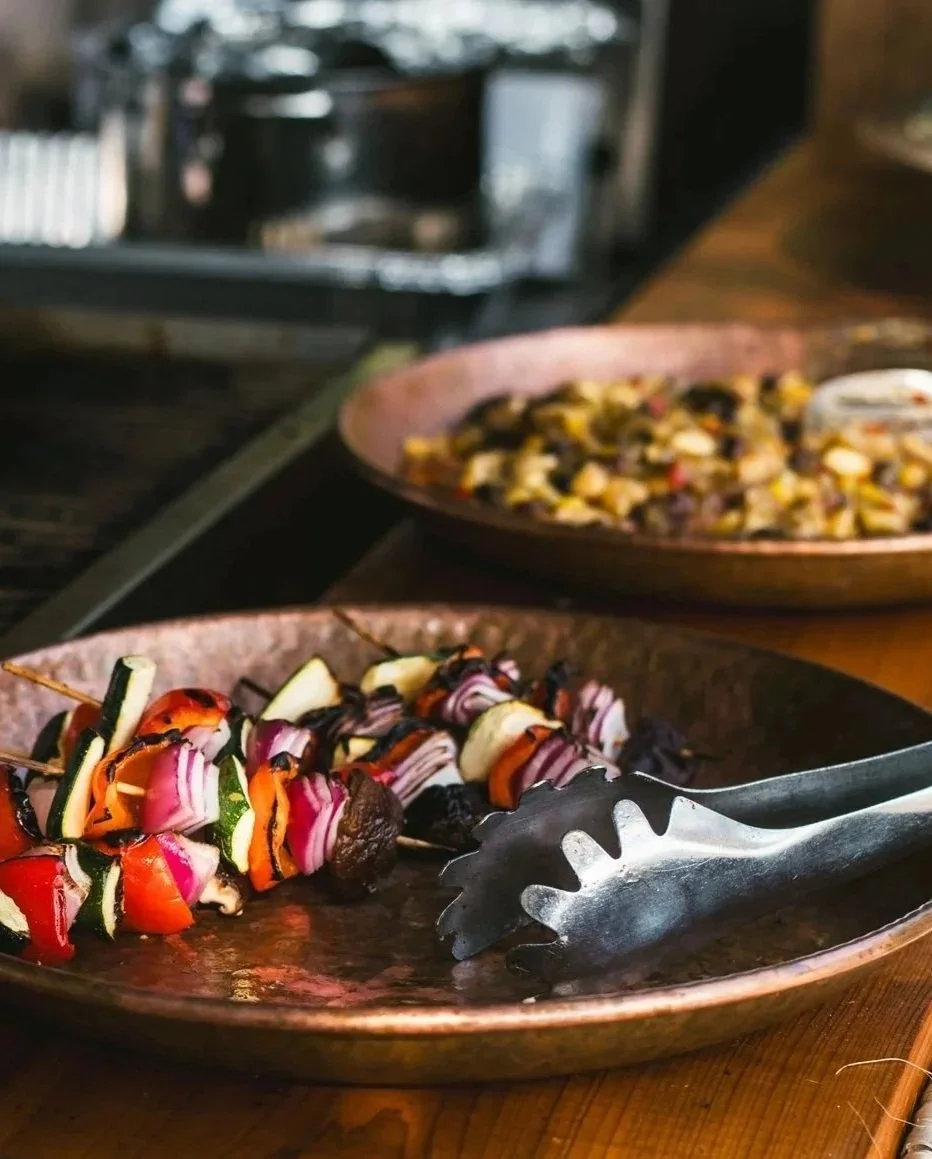 Colorful vegetable skewers on a copper plate with tongs, and a bowl of a mixed bean salad in the background, on a wooden table.