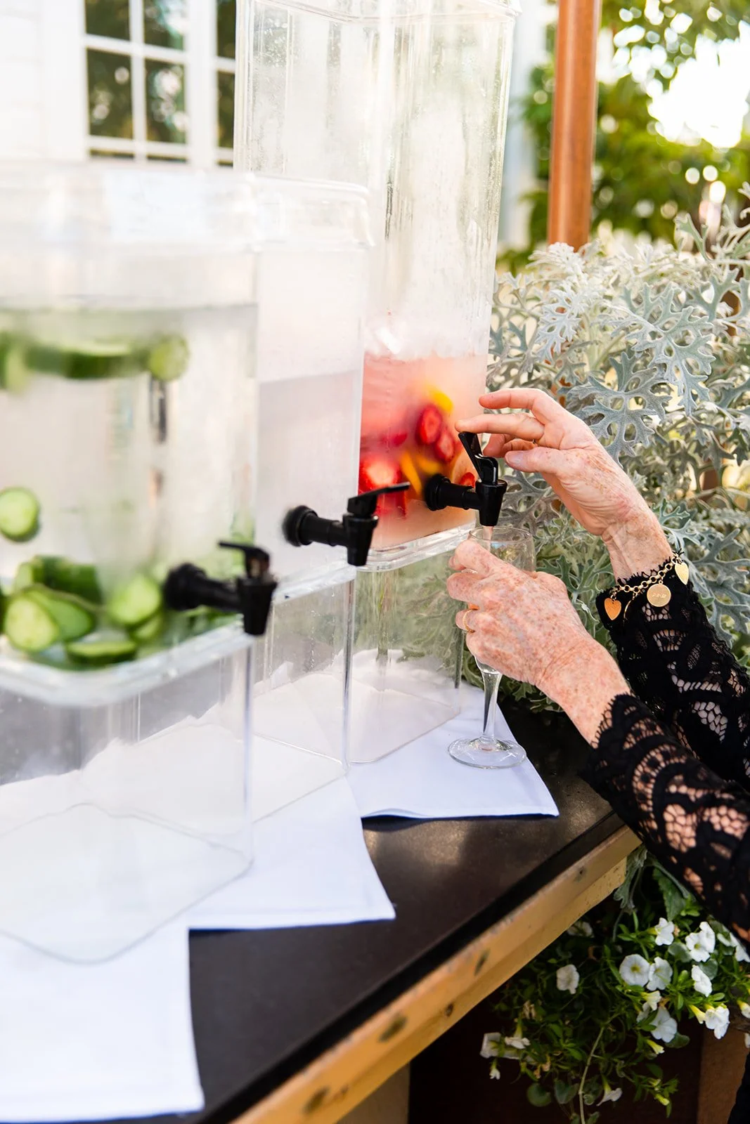 An elderly person with lace long-sleeve black top fills a glass from a fruit-infused water dispenser, which contains strawberries and citrus slices. Two similar water dispensers with cucumbers and another with strawberries are visible on a table outdoors.