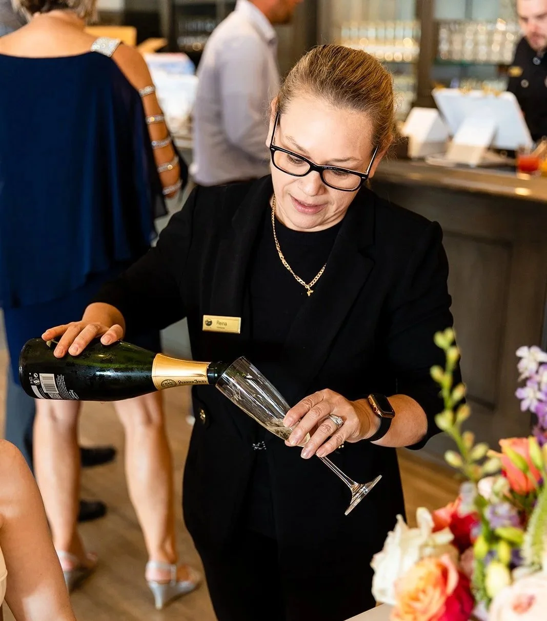 A woman with glasses and a name tag pouring champagne into a flute at a celebration or event.