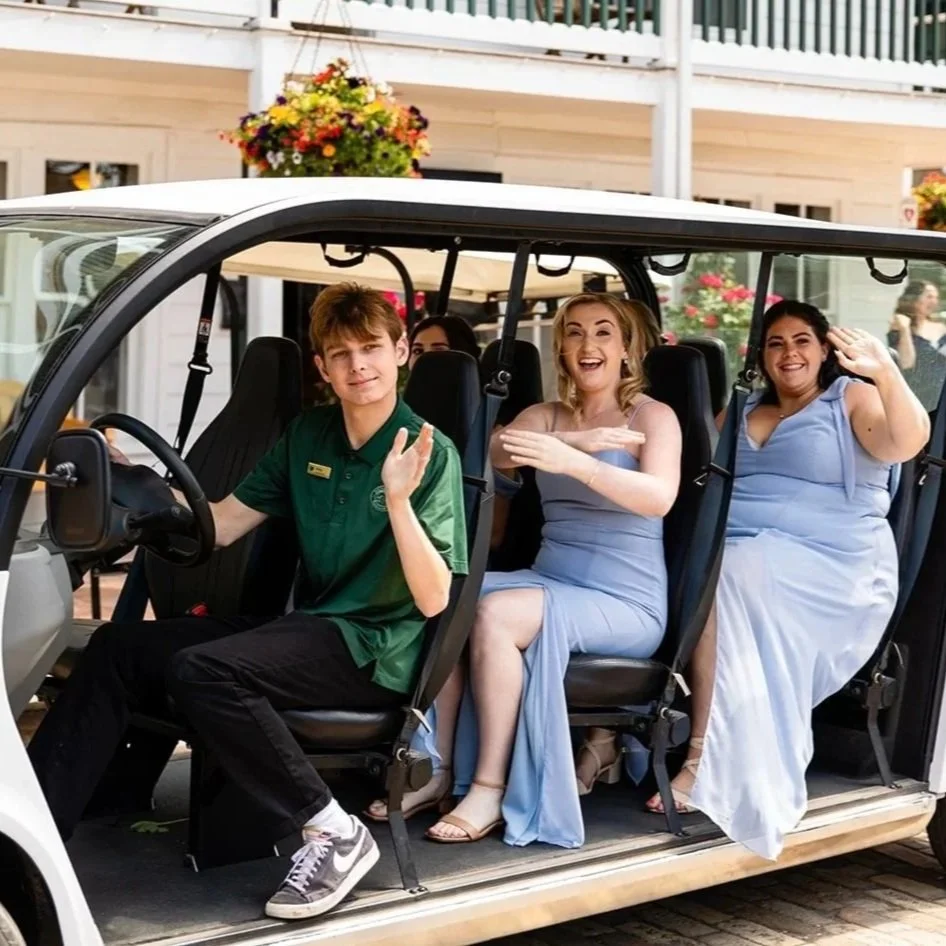 People in fancy dresses riding a golf cart, waving and smiling