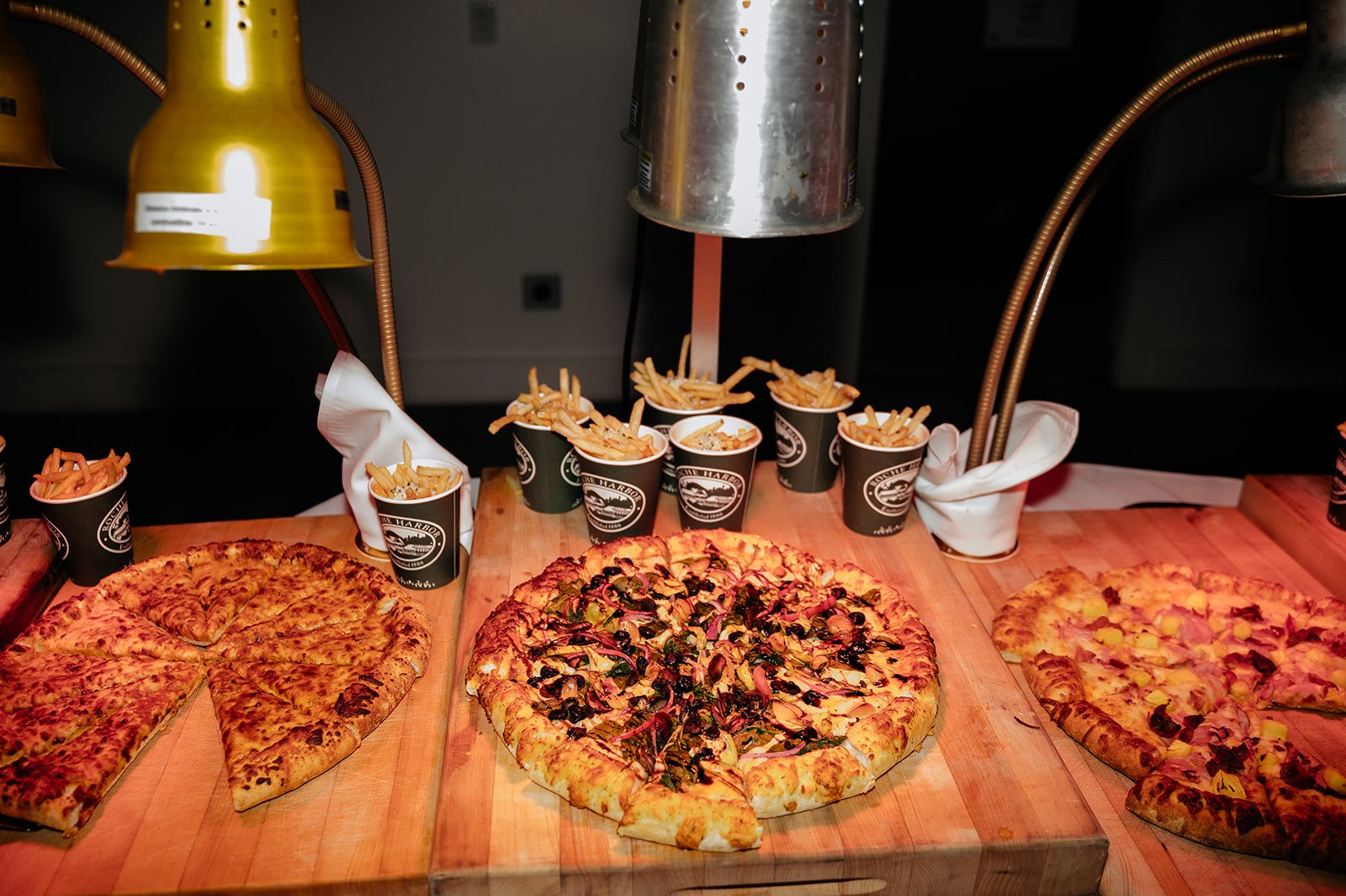 Pizzas and French fries on a wooden table with heat lamps overhead.