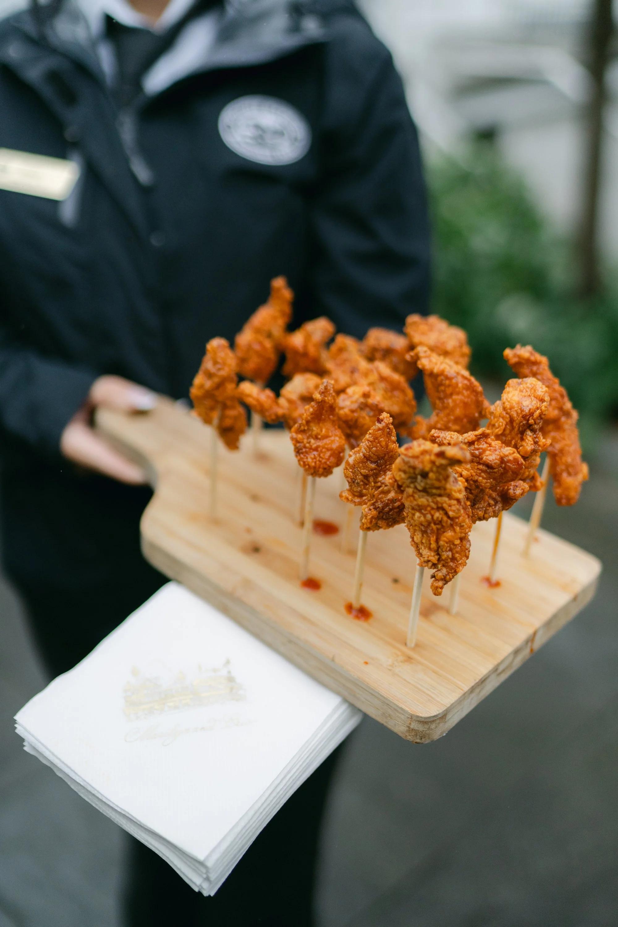 Person holding a wooden platter with six skewers of fried chicken wings, covered in breading and sauce, with napkins underneath.