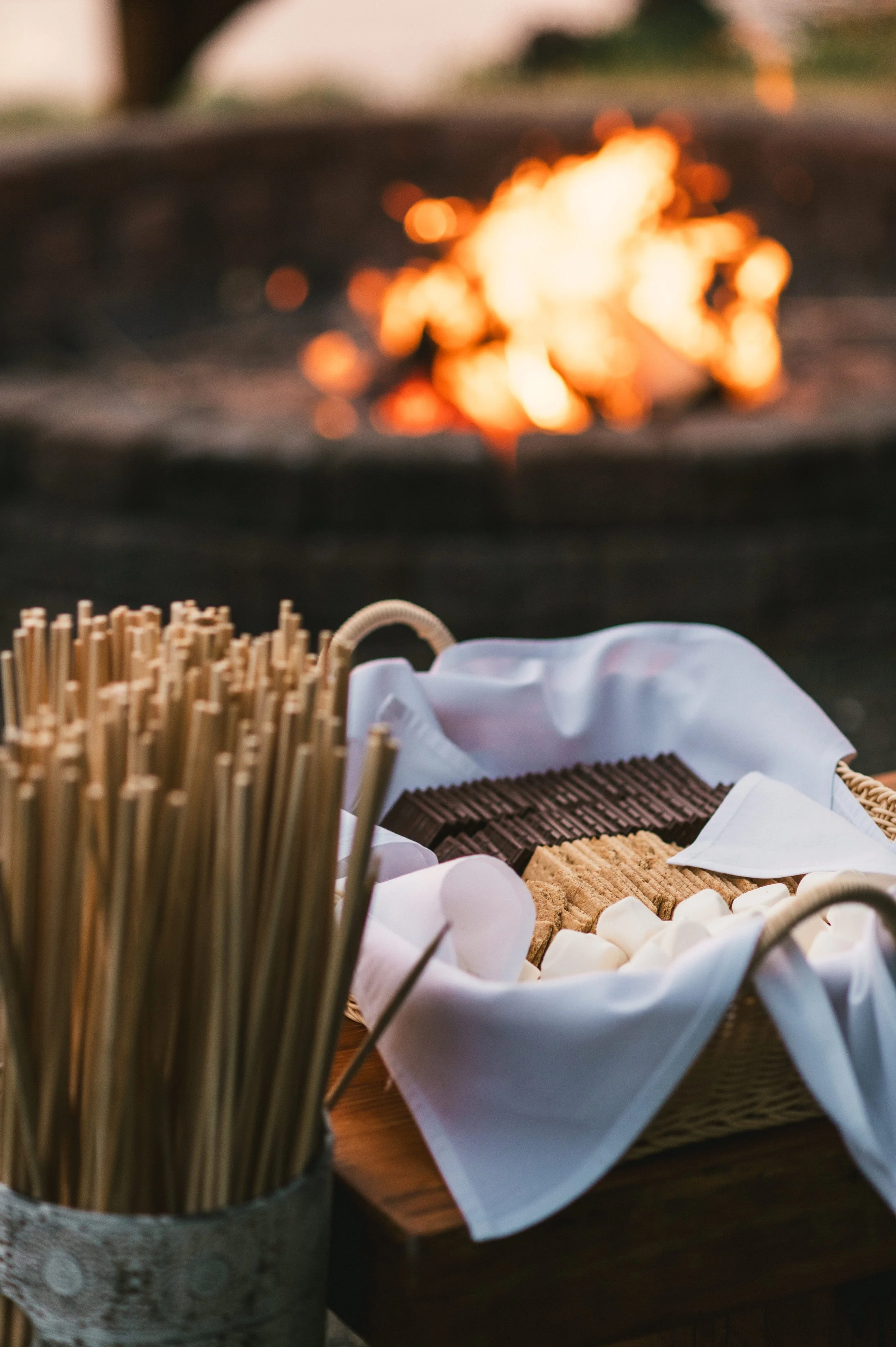 Basket of assorted chocolate and graham crackers with a grouping of bamboo sticks in a container, with a glowing fire in the background.