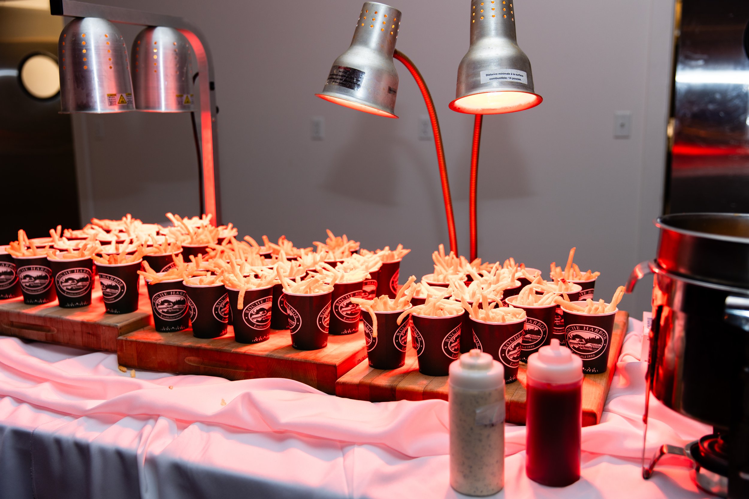Tray of French fries served in black cups on a wooden board at a buffet table with lamps overhead and condiments in bottles in front.