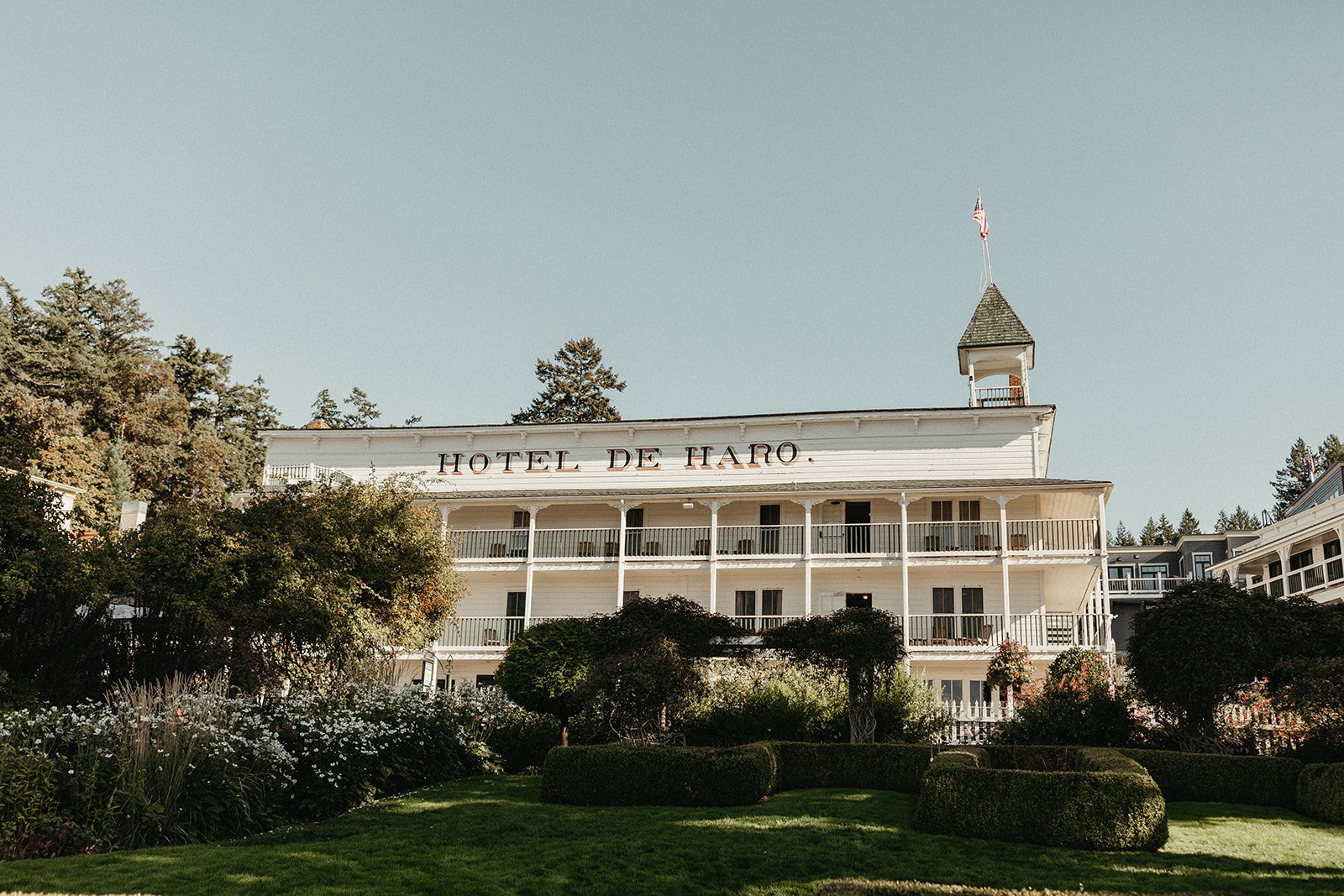 White multi-story building labeled 'Hotel de Haro' with green landscaped garden in front, surrounded by trees, with a tower and American flag on top, under a clear blue sky.