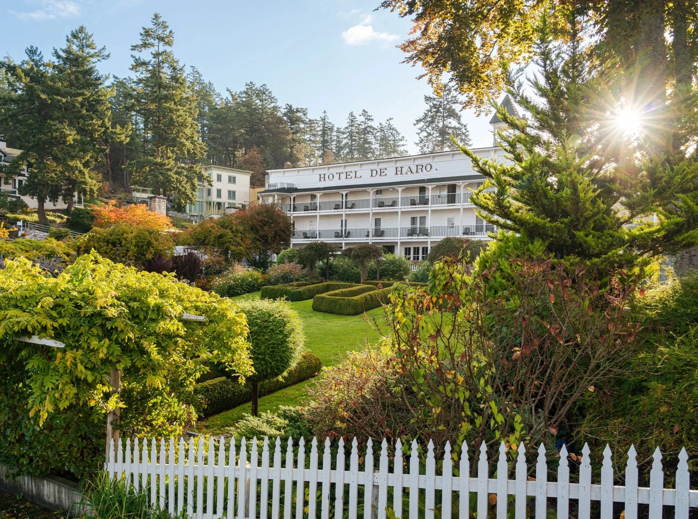 The image shows a lush green garden with trimmed bushes, trees, and a white picket fence in the foreground. In the background, there are multi-story buildings, including a prominent white building labeled 'Hotel de Haro,' surrounded by tall pine trees under a sunny sky.