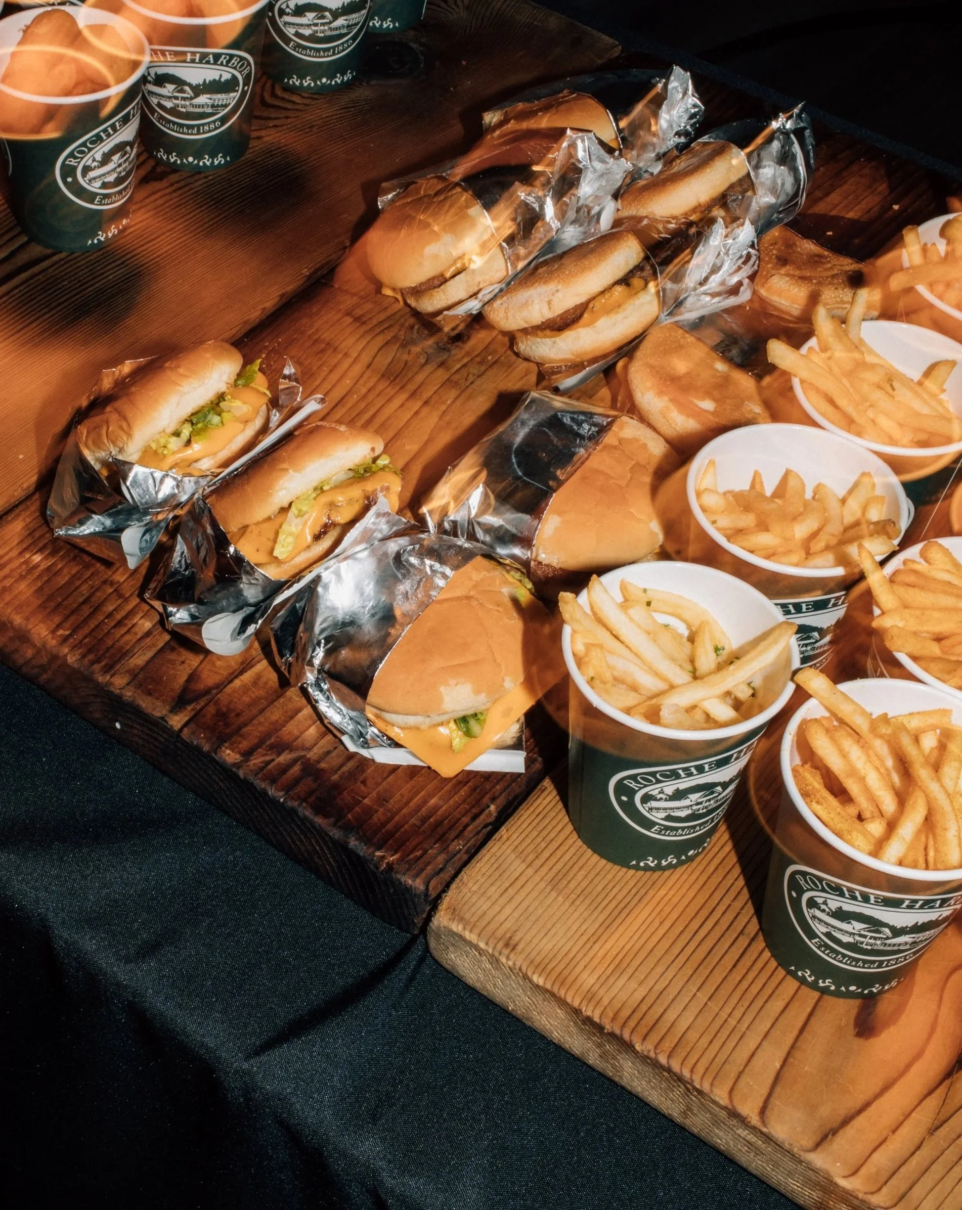 An assortment of wrapped hot dogs and cheeseburgers on a wooden serving board, with cups of French fries in branded paper cups.