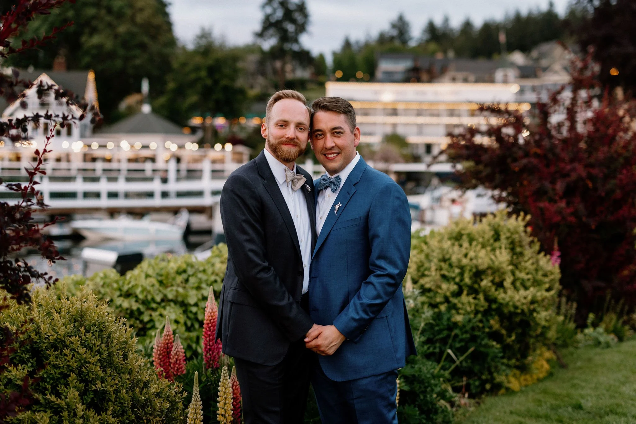 Two men in suits holding hands and smiling at the camera outdoors, with a marina and boats in the background, surrounded by greenery and flowering plants.