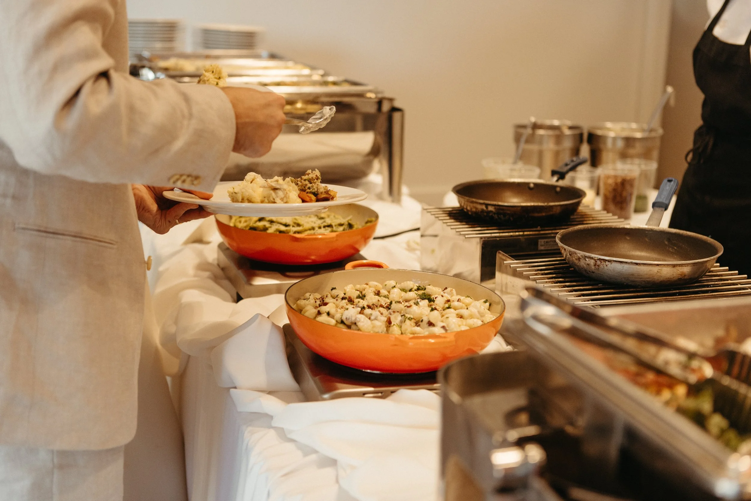 Person serving food at a buffet-style table with dishes including cauliflower, pasta, and other side dishes.
