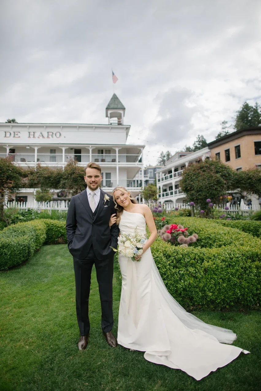 Bride and groom standing together on a garden lawn in wedding attire smiling, with a large white house and cloudy sky in the background.
