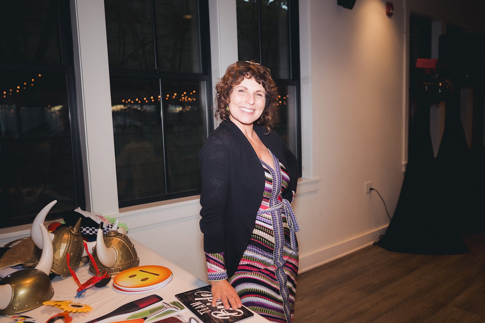 A woman with curly brown hair smiling and standing indoors near a table with various costume accessories, including horns, masks, and emojis, in front of a window at night.