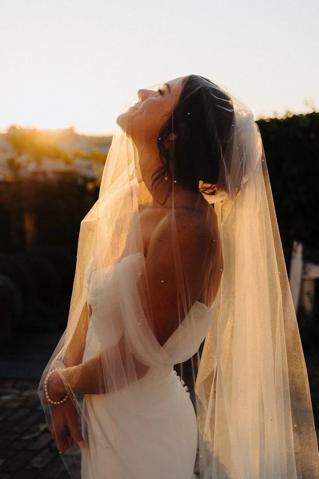 A bride standing outdoors at sunset, smiling and looking upwards, wearing a wedding dress and veil.