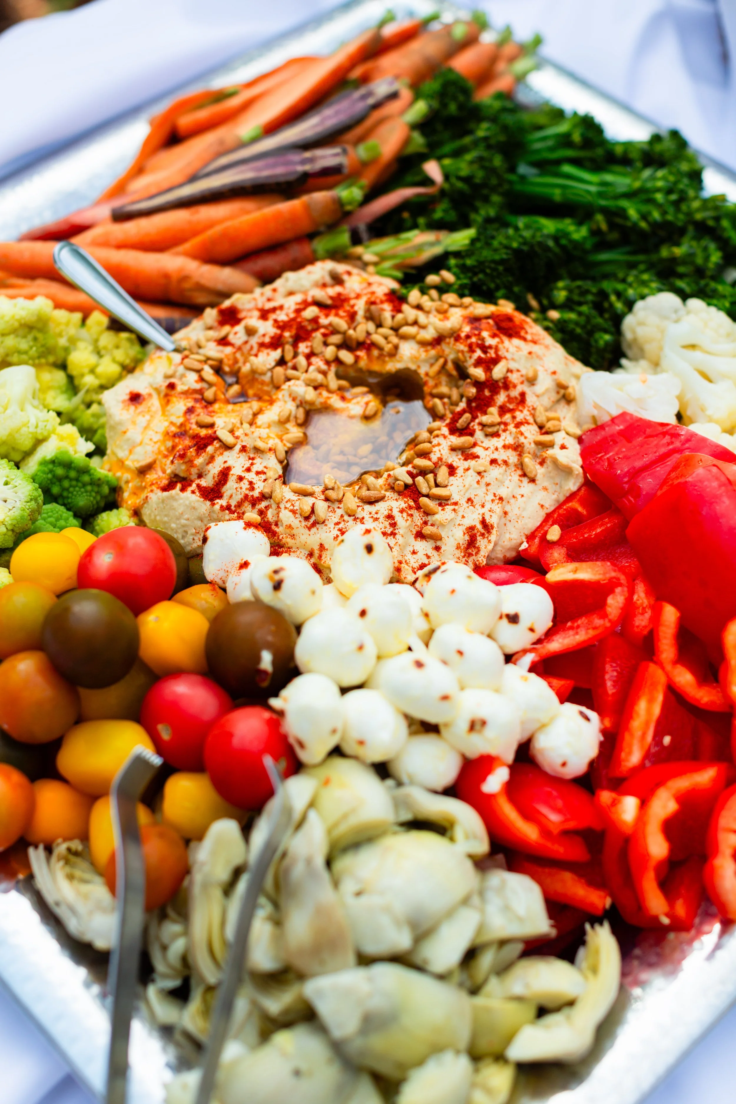 A large tray of assorted raw vegetables and a bowl of hummus with chopped nuts and paprika.