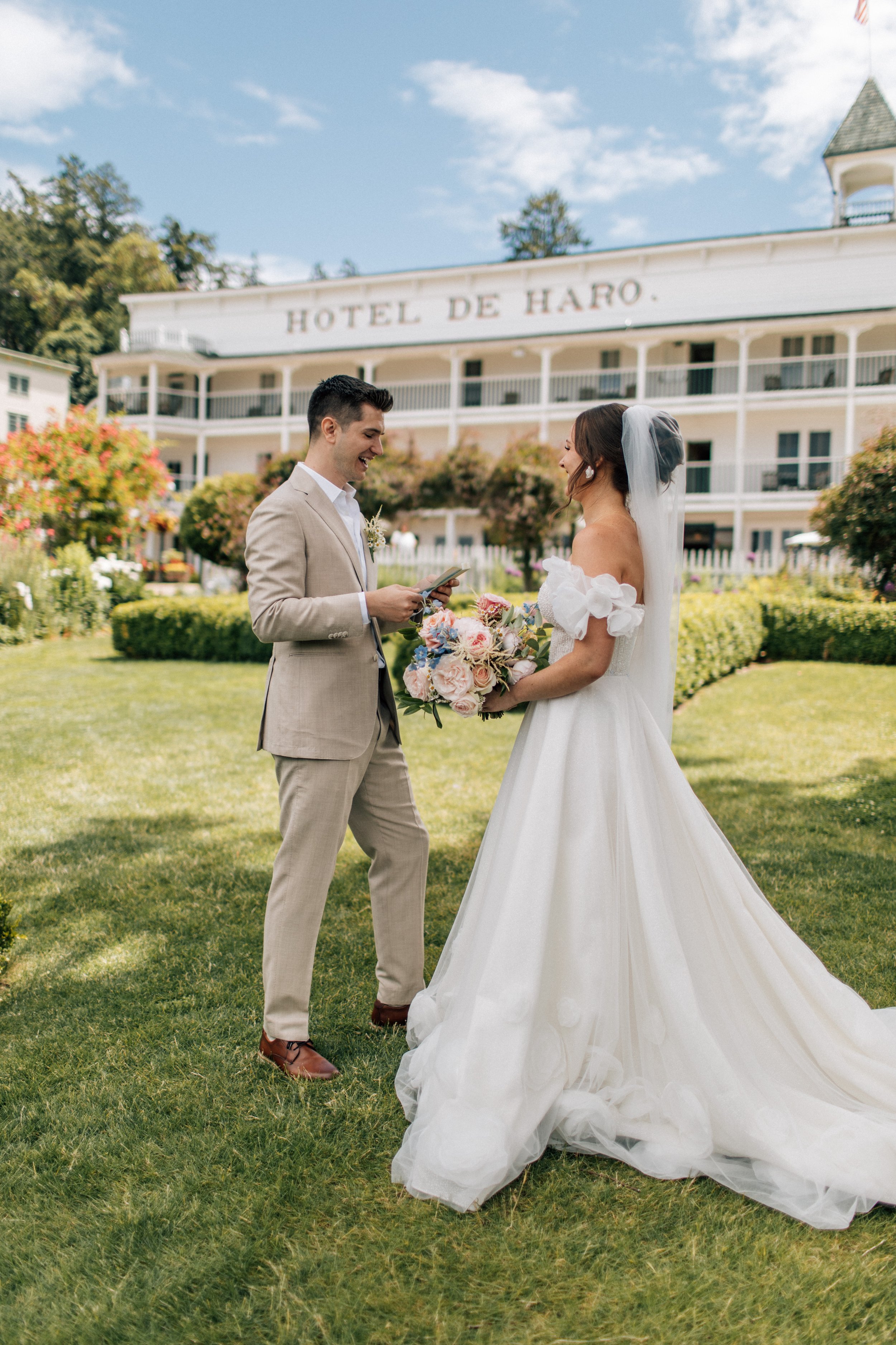 A bride and groom stand outdoors on a grassy lawn with a hotel building in the background. The groom is reading from a small book, and the bride is holding a bouquet of flowers, both smiling.