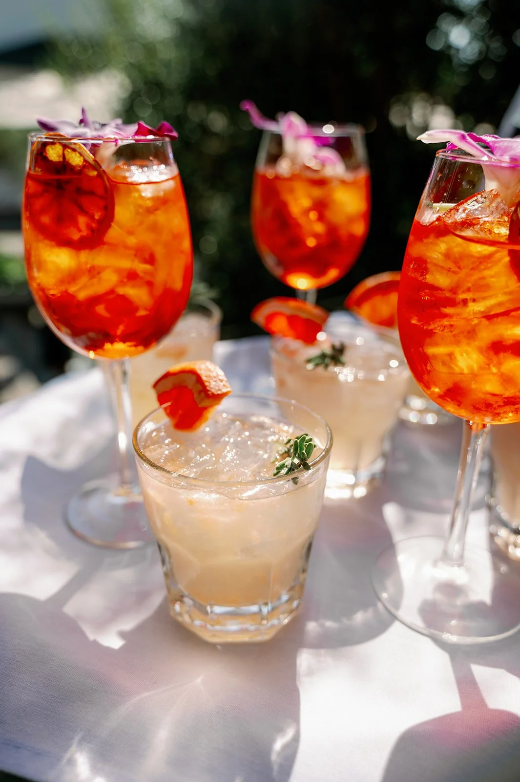 Several glasses of orange-colored cocktails with ice, garnished with slices of citrus fruit and edible flowers on a white tablecloth outdoors.