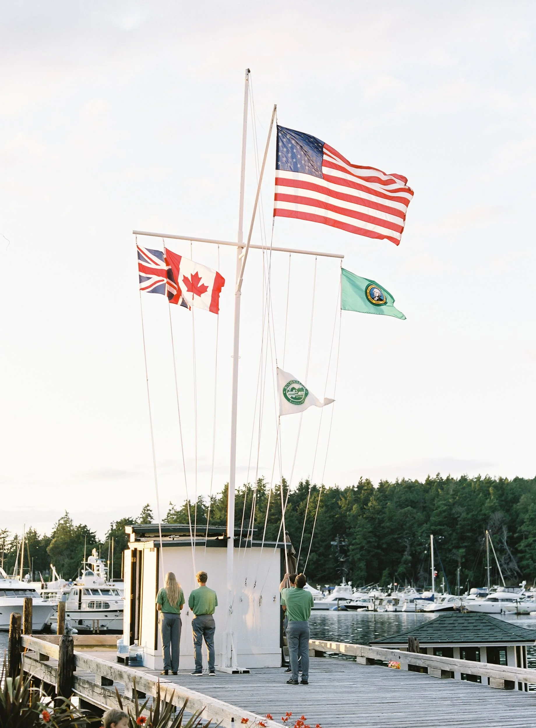 People in green shirts standing on a dock, lowering flagpoles with flags, including the American flag, at a marina during daytime.