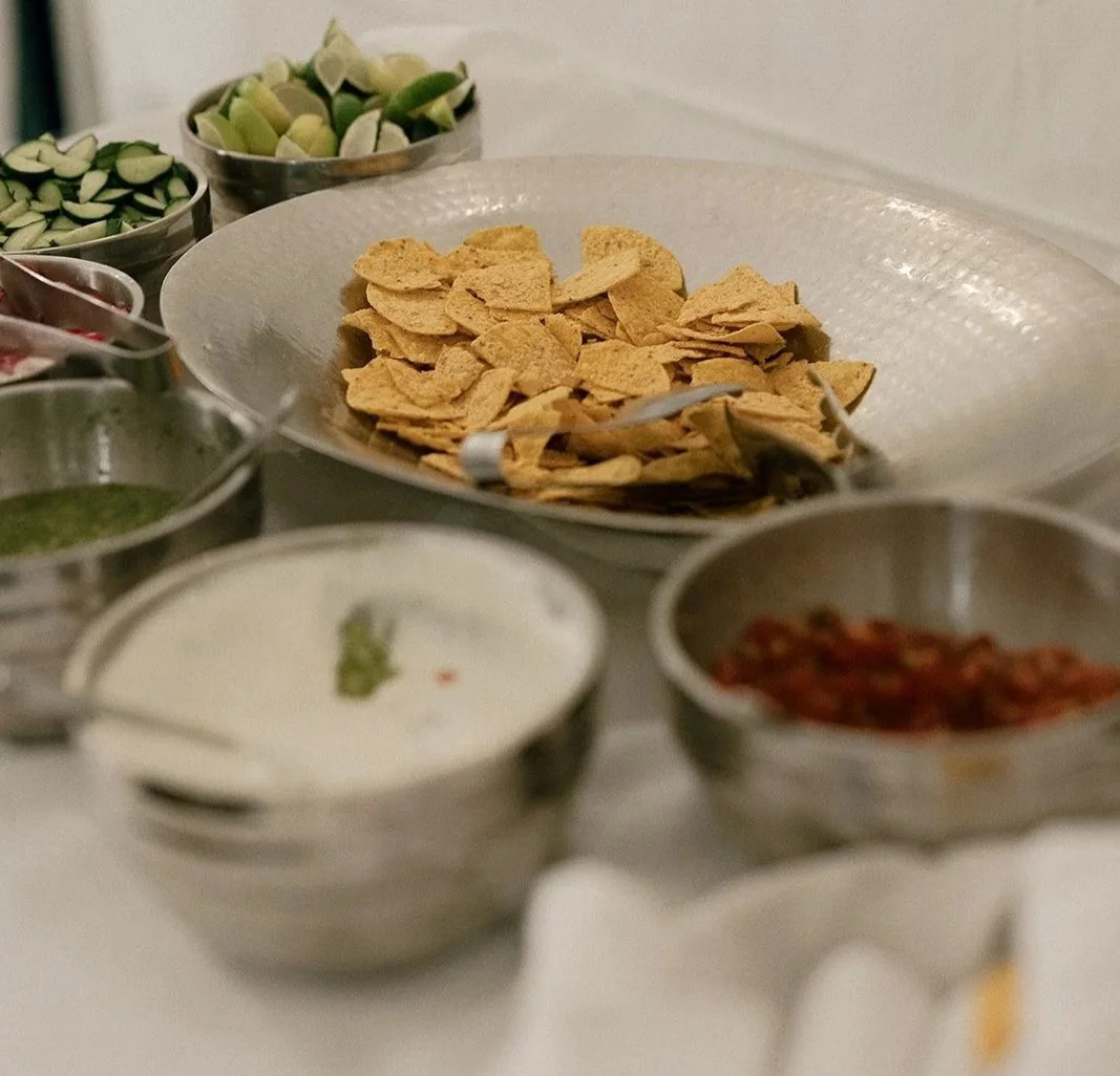 A buffet table with bowls of snacks and condiments, including pita chips, sliced vegetables, sauces, and toppings.