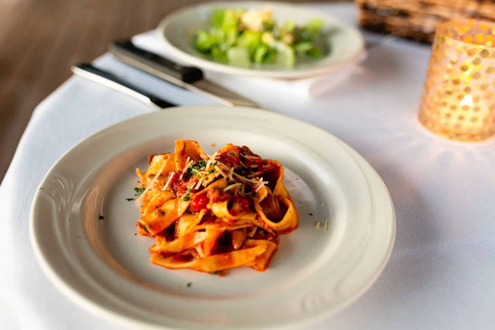A plate of pasta with tomato sauce, garnished with chopped herbs and shredded cheese, on a white tablecloth, with a bowl of salad and a lit candle in the background.