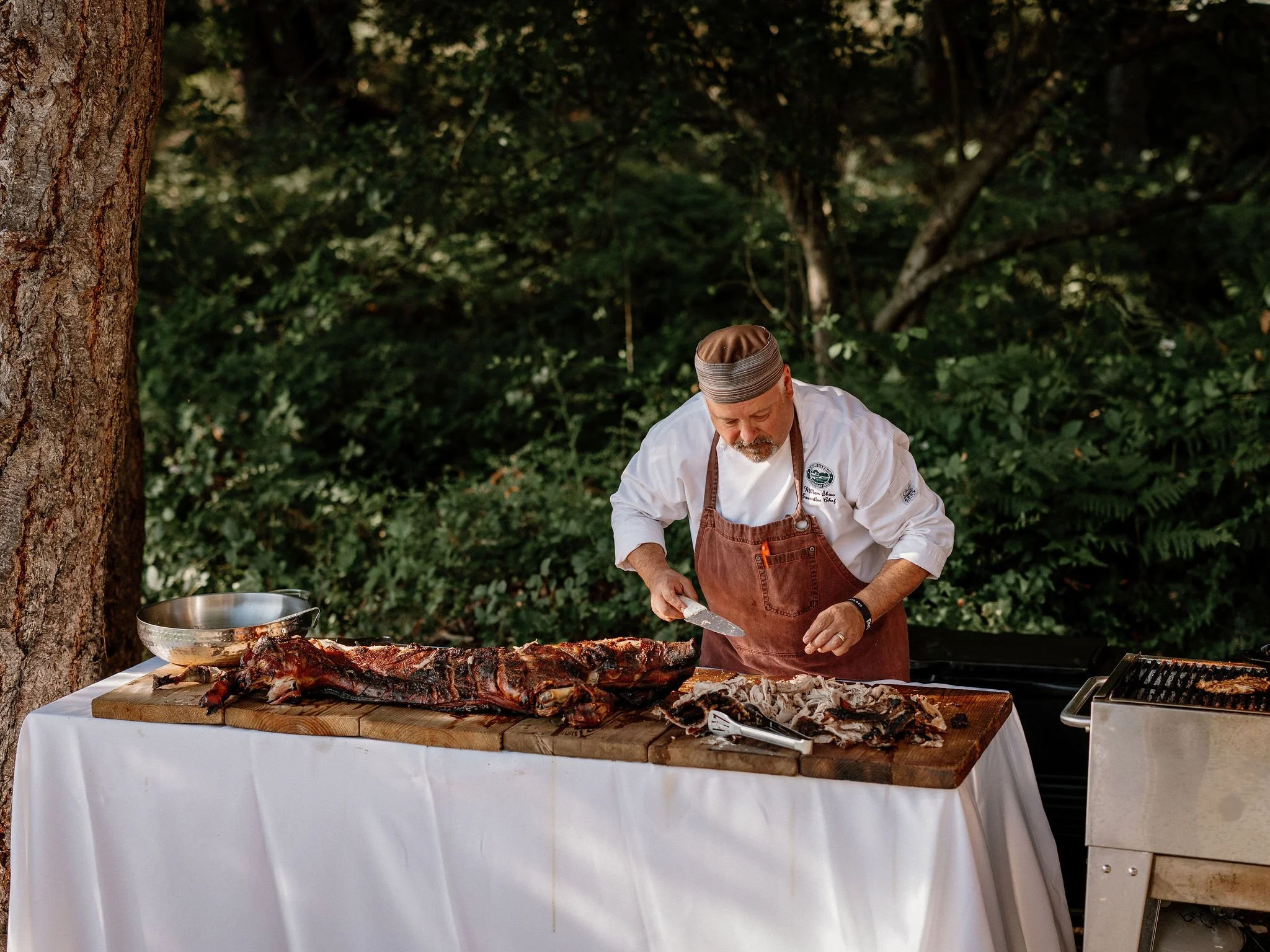 Chef in outdoor setting carving cooked meat on a wooden cutting board, with a grill visible on the right.
