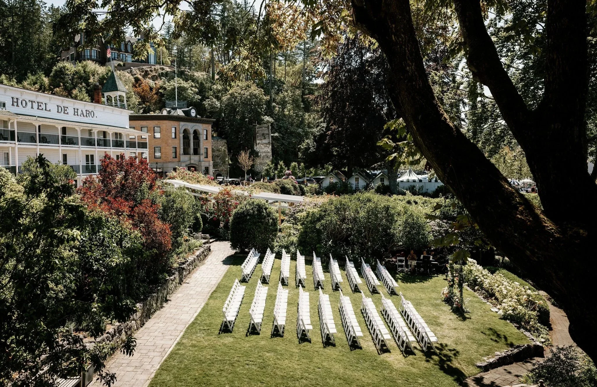 Outdoor garden with rows of white chairs set up for an event, surrounded by greenery and trees, overlooking a hotel building in the background.