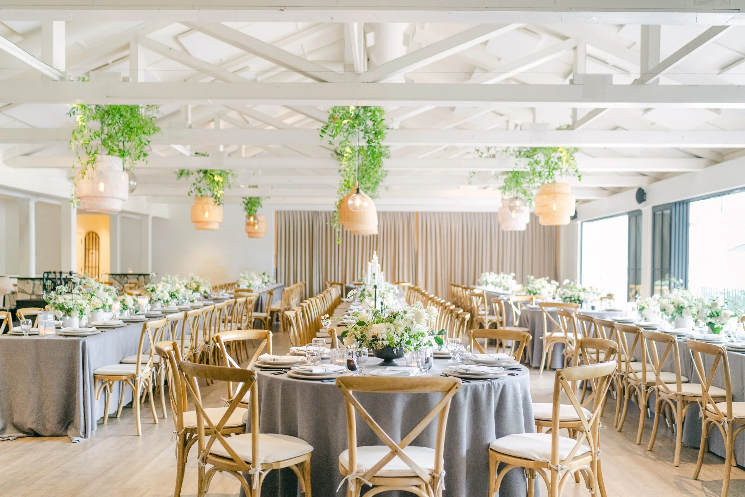 Elegant banquet hall with round tables covered in gray tablecloths, decorated with white floral centerpieces. Wooden cross-back chairs surround the tables. Hanging wicker pendant lights and indoor plants decorate the bright, modern space with large windows and beige curtains.