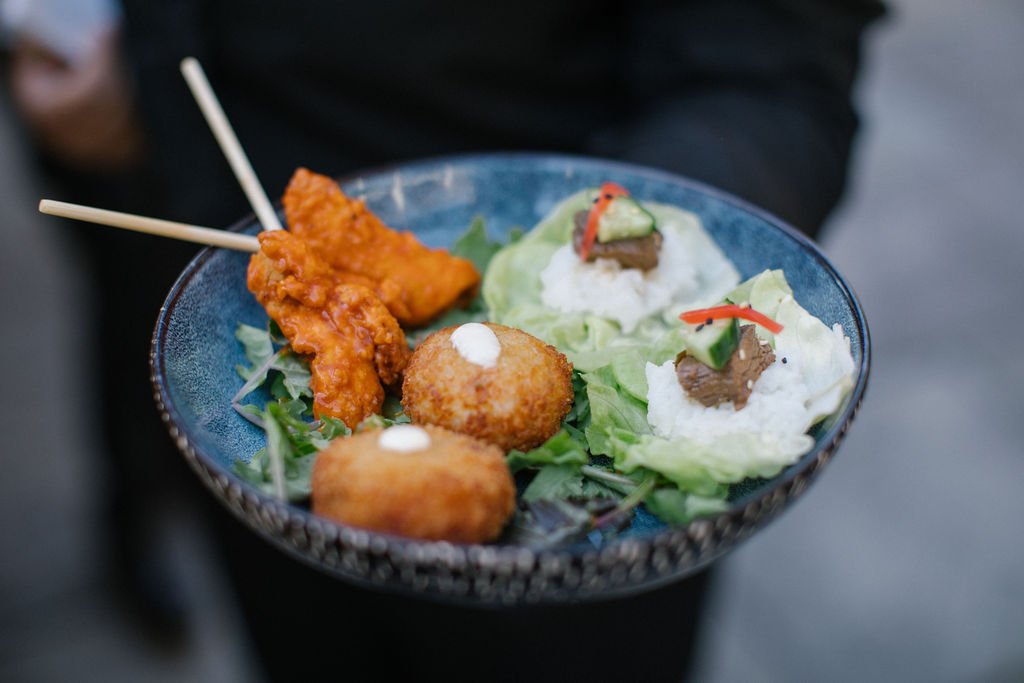 A blue bowl containing chicken skewers, a fried ball, and two lettuce cups topped with sauce and garnishes, on a person's lap.