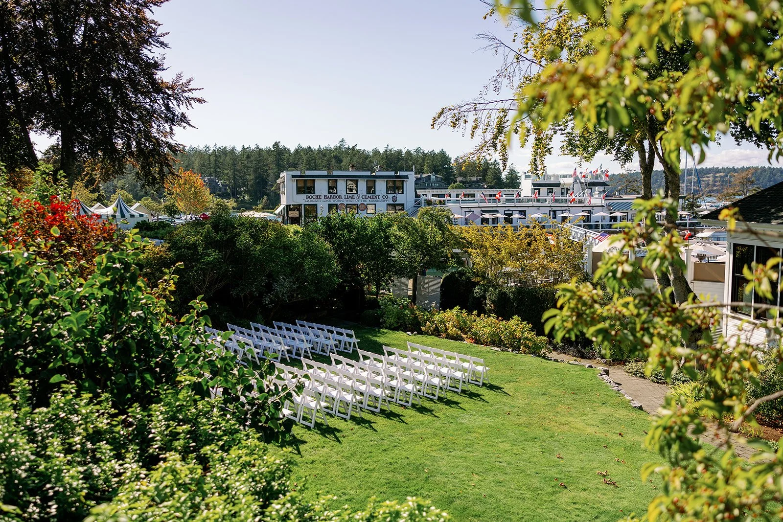 Outdoor wedding ceremony setup with rows of white chairs on a grassy lawn, surrounded by trees and bushes, with a marina and boats in the background.