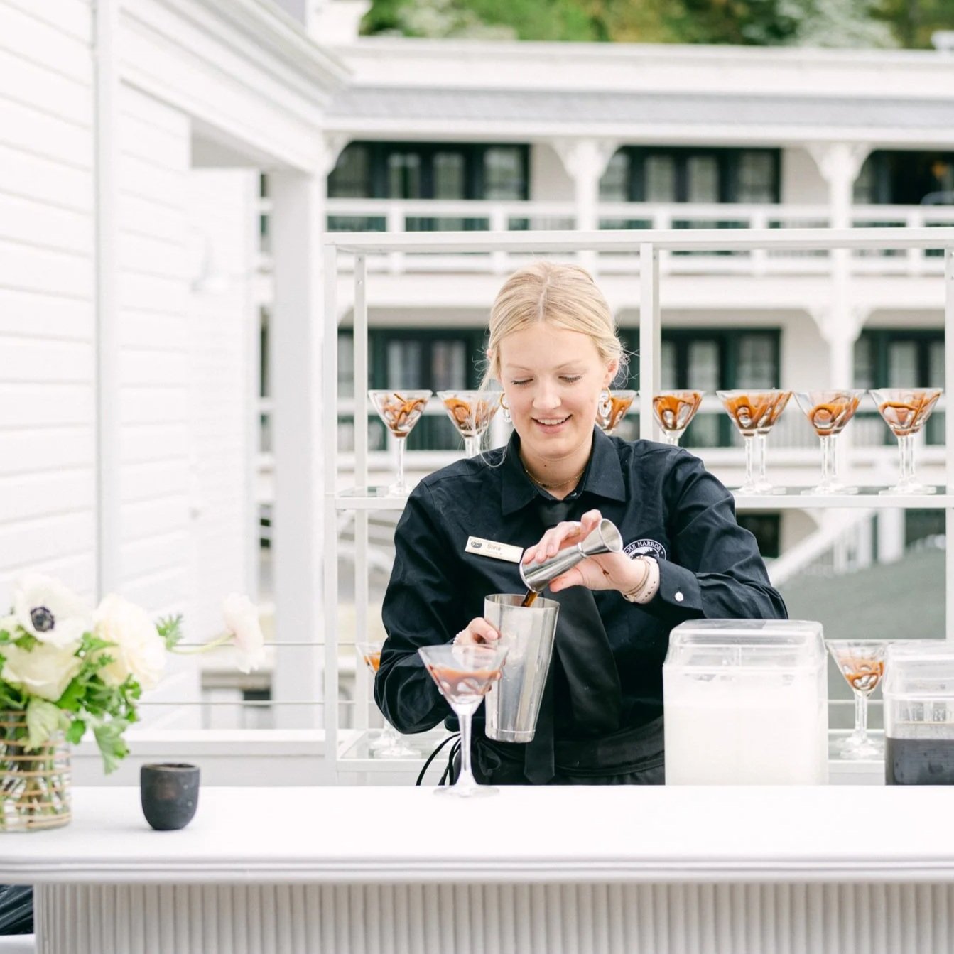 A woman in black uniform pouring a drink at an outdoor bar with glasses of martinis and a flower arrangement nearby.
