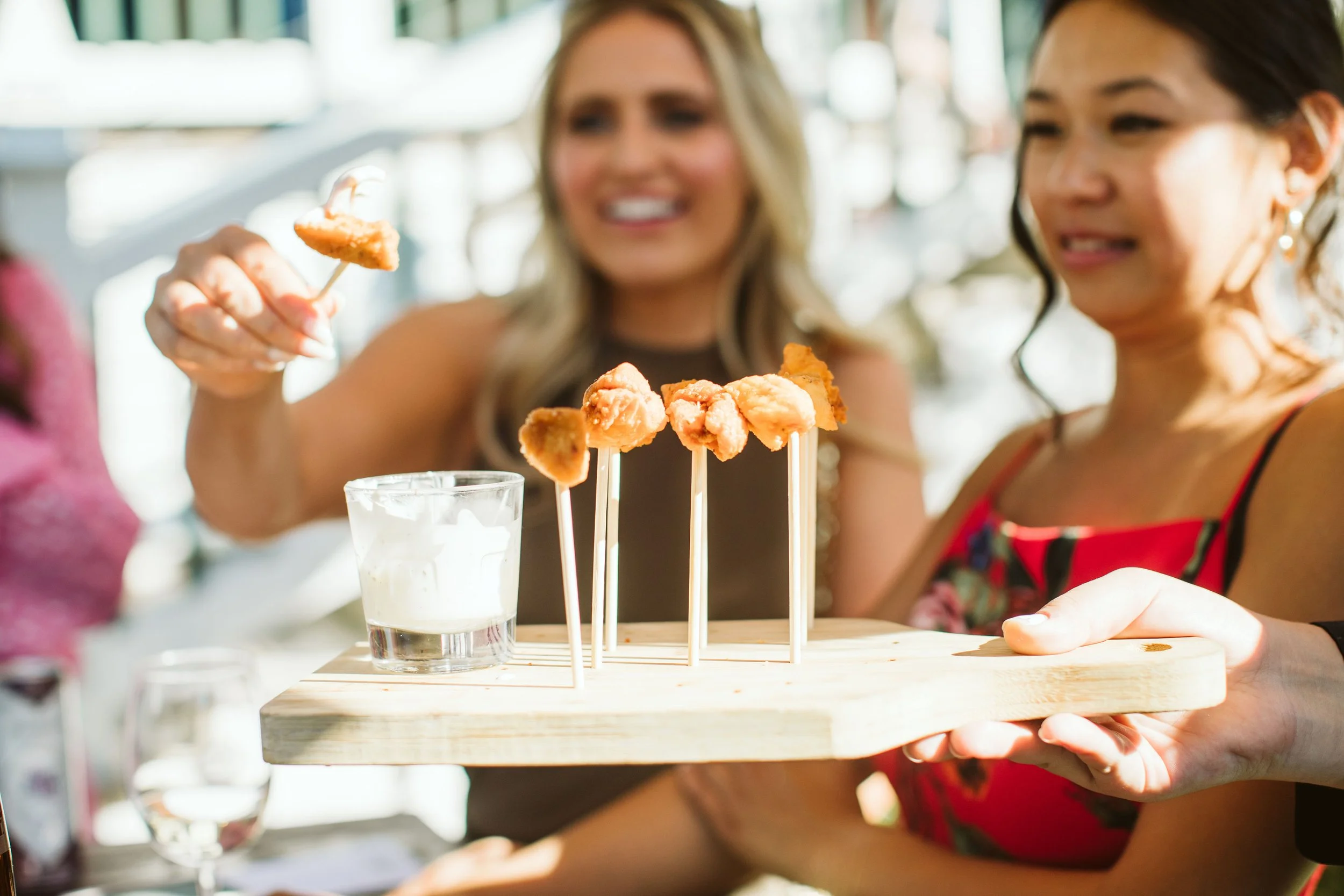 Two women at a restaurant table with a wooden platter of fried chicken pieces on sticks, one woman is holding a piece over the platter, and there is a glass of water in front of her.