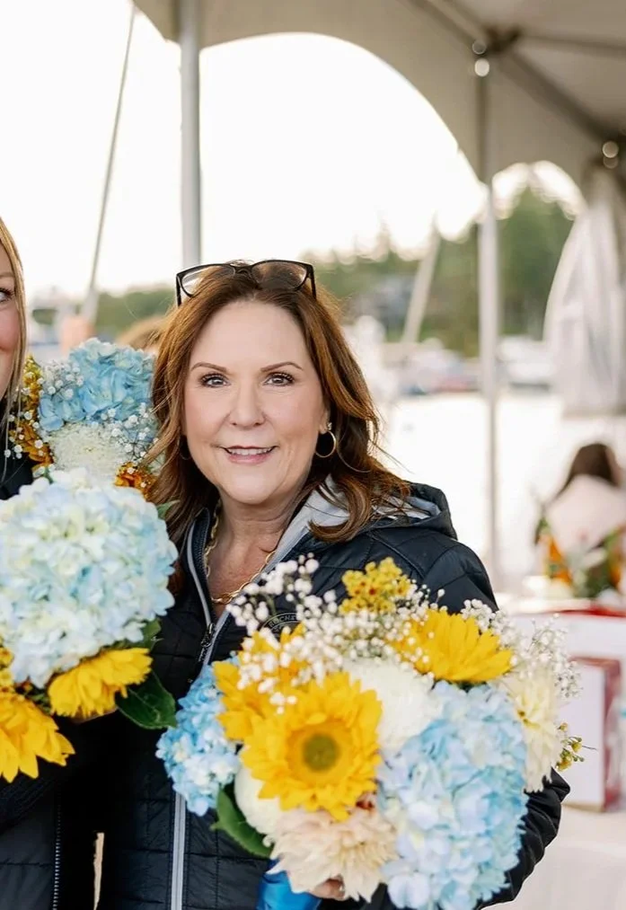 A woman with brown hair, smiling, holding a bouquet of yellow, blue, and white flowers, standing under a tent outdoors.