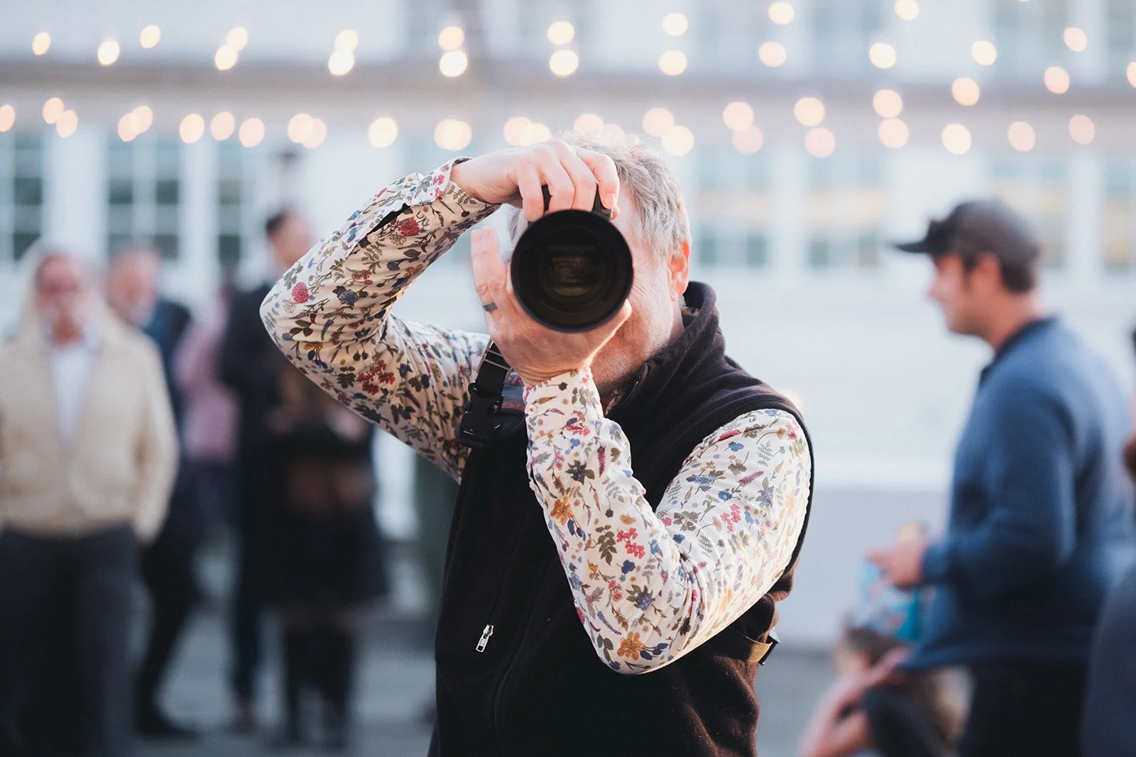A man taking a photograph with a camera at an outdoor event, with blurred people in the background and string lights overhead.
