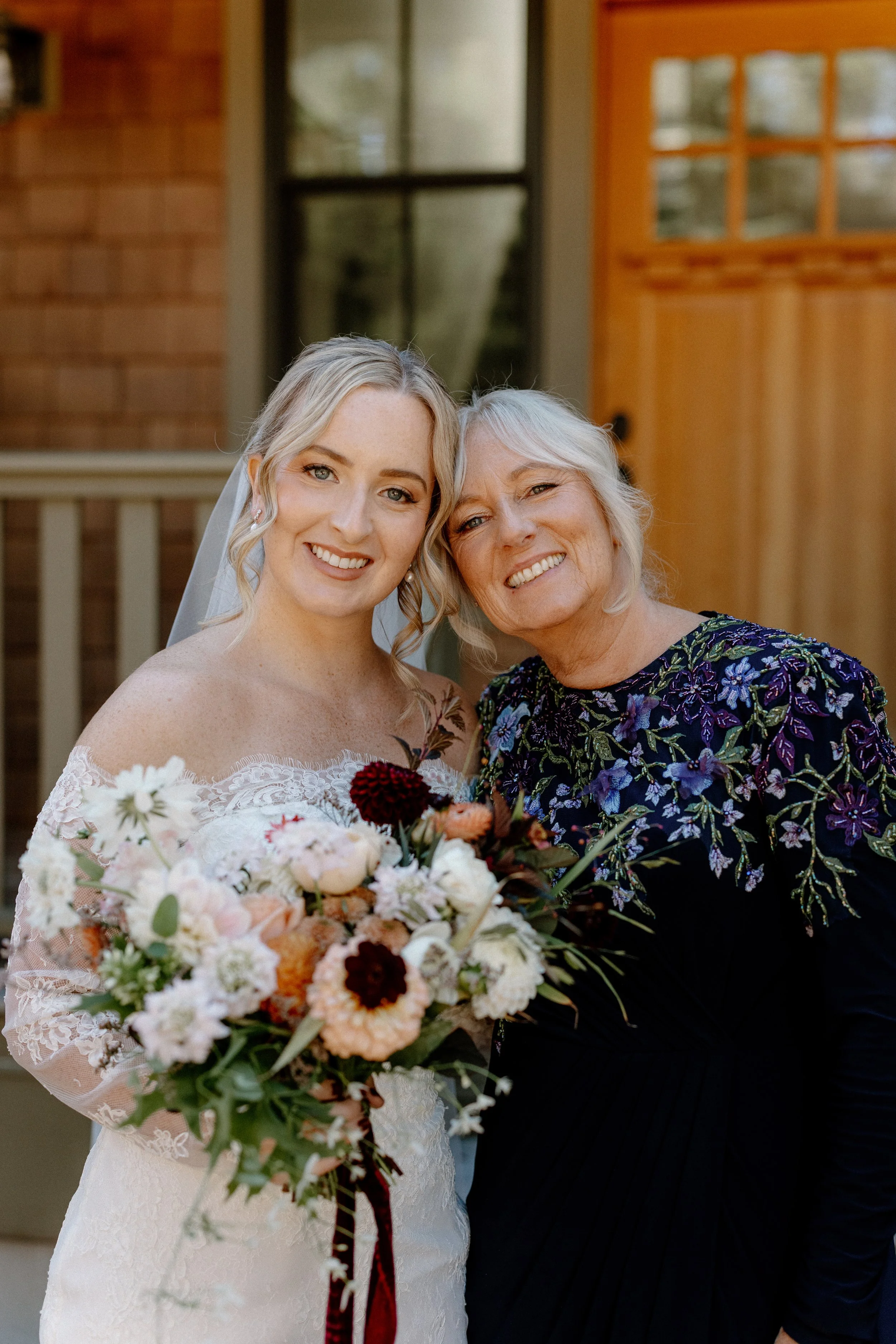 A bride in a white lace wedding dress holding a bouquet of flowers, standing next to an older woman with short gray hair wearing a dark dress with floral embroidery, both smiling outside in front of a brick and wooden house.