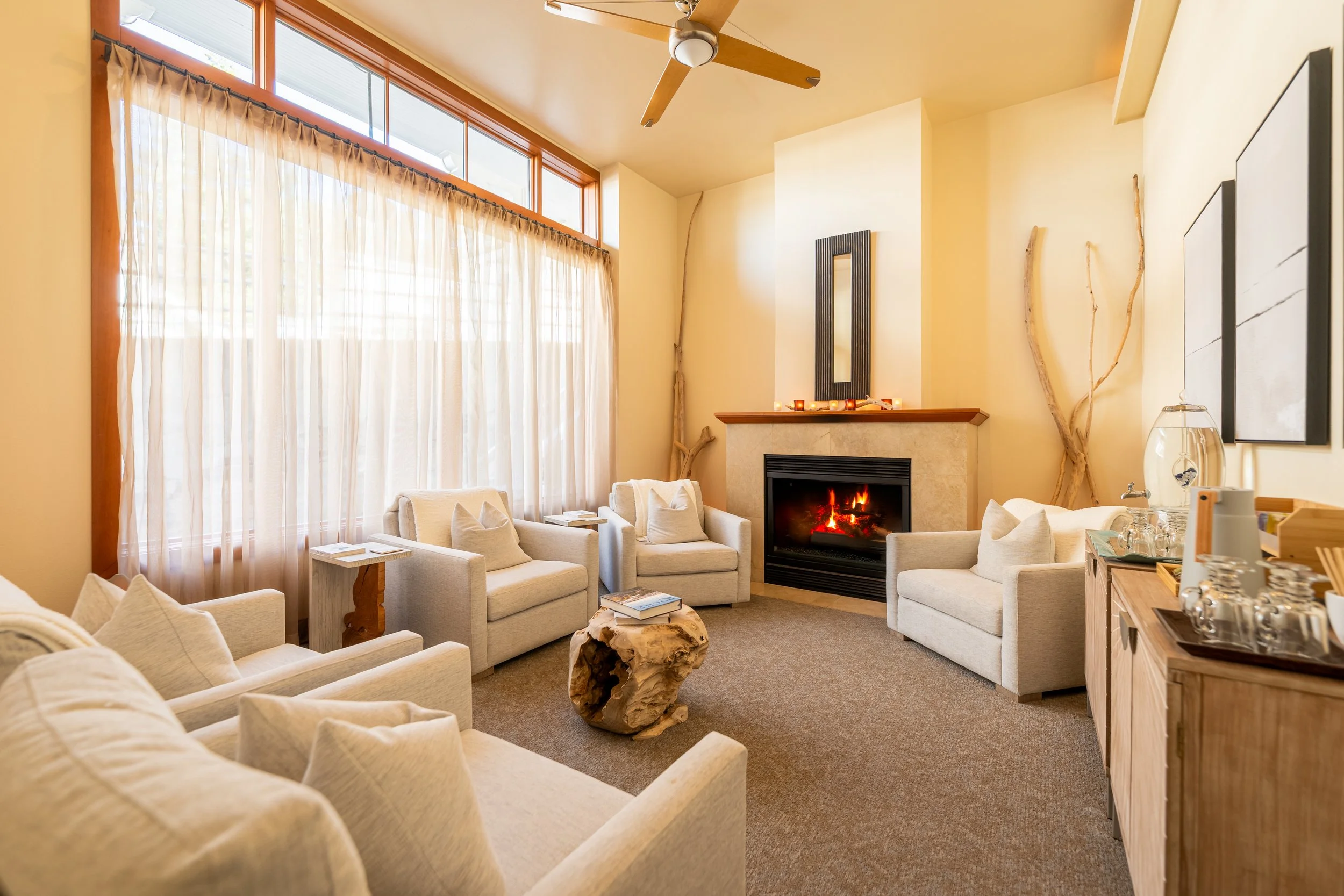 A cozy living room with cream-colored sofas and armchairs arranged around a fireplace with a lit fire. Large windows with sheer curtains let in natural light. There are decorative sticks in the corners, a wooden coffee table, a ceiling fan, and a wooden sideboard with glasses and a water dispenser.