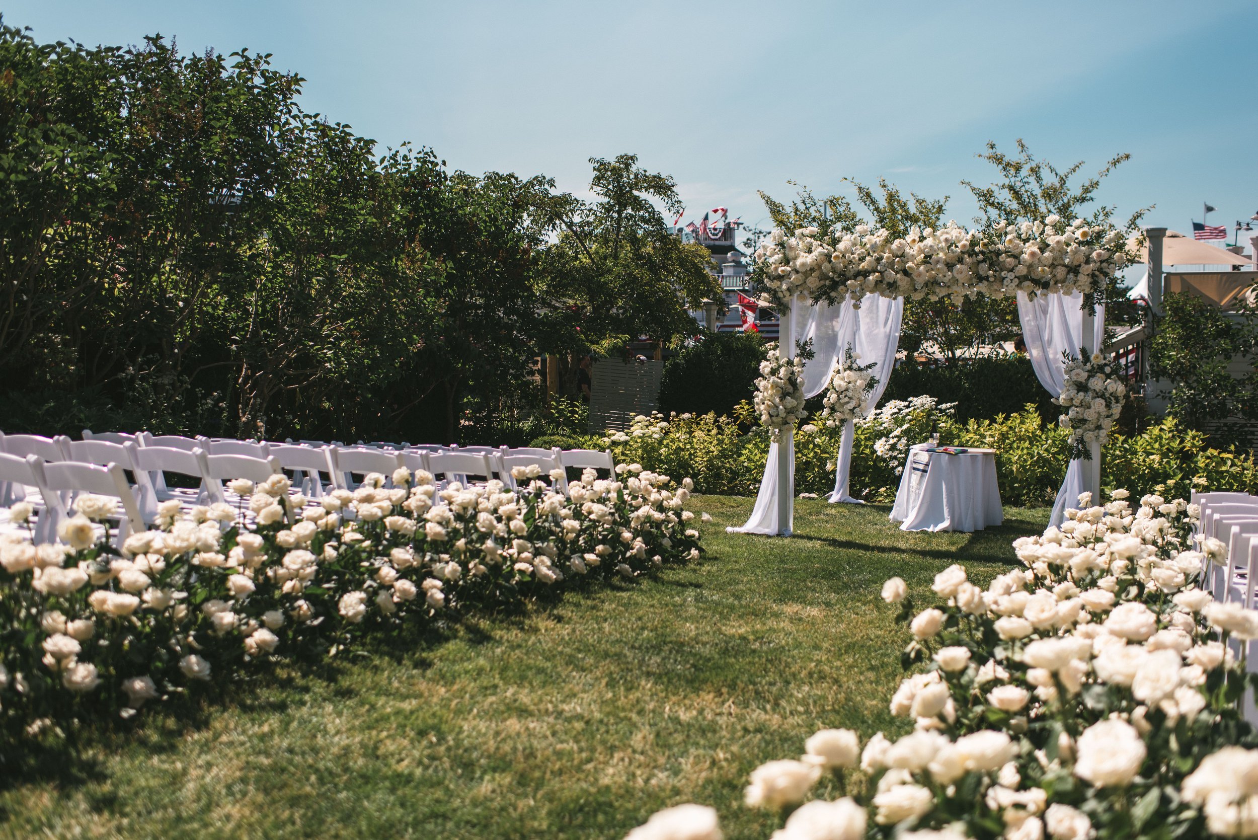 Outdoor wedding ceremony setup with white chairs, floral arrangements, and a decorated arch on a grassy area, with trees and a blue sky in the background.