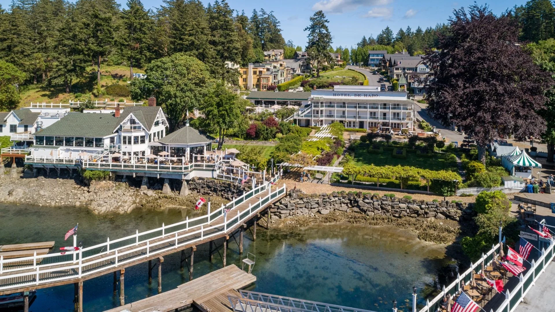 A scenic view of a lakeside restaurant with white railings, outdoor seating, and American flags, overlooking a calm water body with rocky edges, surrounded by lush green trees and high-end houses on a hillside.