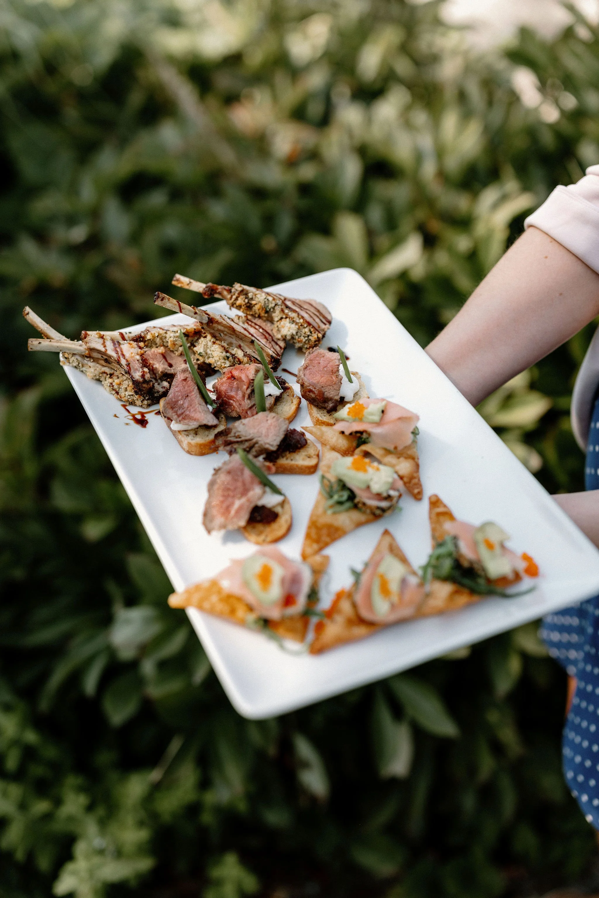 A person holding a white rectangular plate with an assortment of appetizers including lamb chops, small sandwiches, and cheese-topped crackers set outdoors against green foliage.