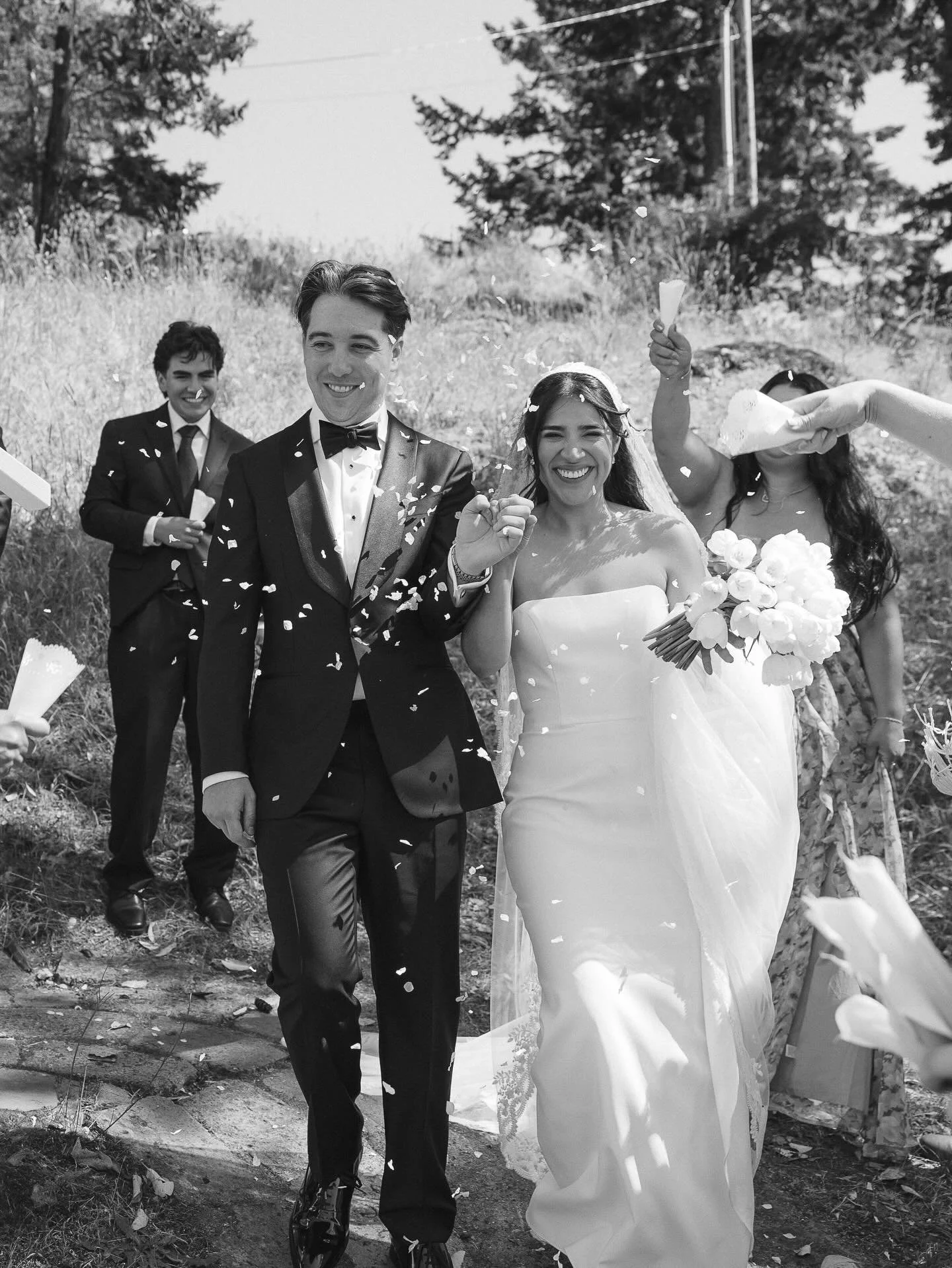 A bride and groom walking outdoors after their wedding, surrounded by well-dressed guests. The bride is holding a bouquet of flowers, and guests are celebrating with confetti.
